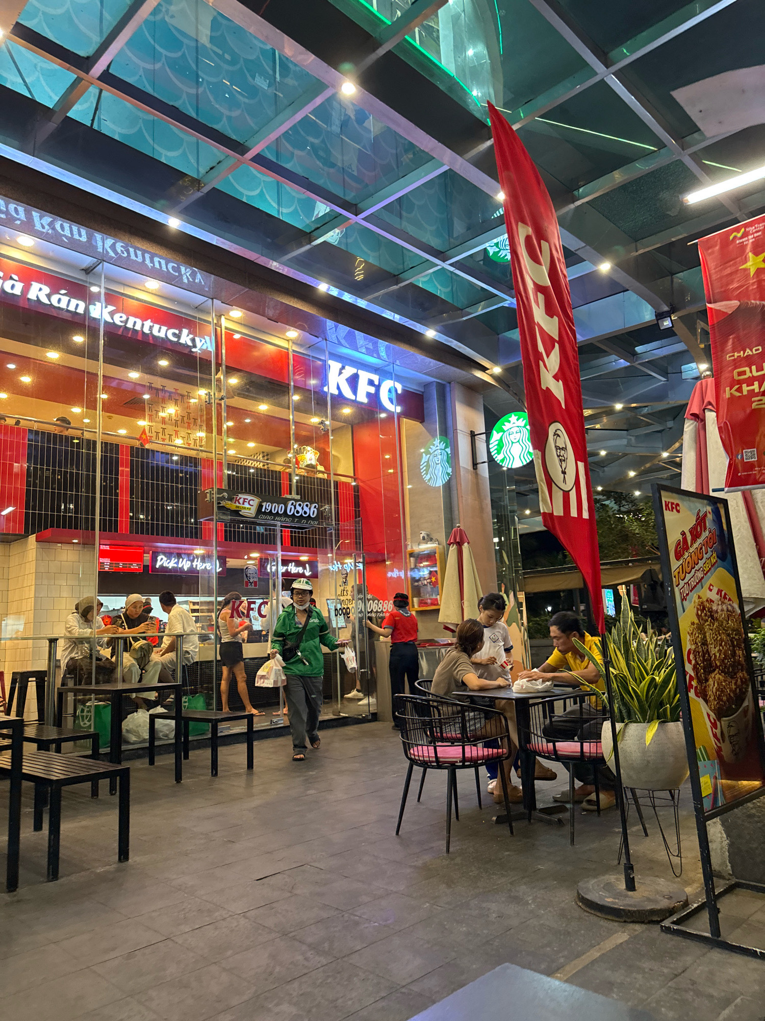 This image shows the exterior of a KFC restaurant in Nha Trang, Vietnam, at night. The restaurant has a large glass facade with the KFC logo and branding visible. Inside, there are several customers seated at tables and others walking around. Outside, there are also some tables and chairs set up, with a few people sitting and eating. A large red KFC flag is prominently displayed, along with a Starbucks logo on the building. The street is paved and dimly lit, with overhead lighting from the restaurant and streetlights. A man in a green jacket is walking away from the KFC with a takeout bag in his hand. The overall atmosphere is busy and commercial, typical of a fast-food establishment in a city.