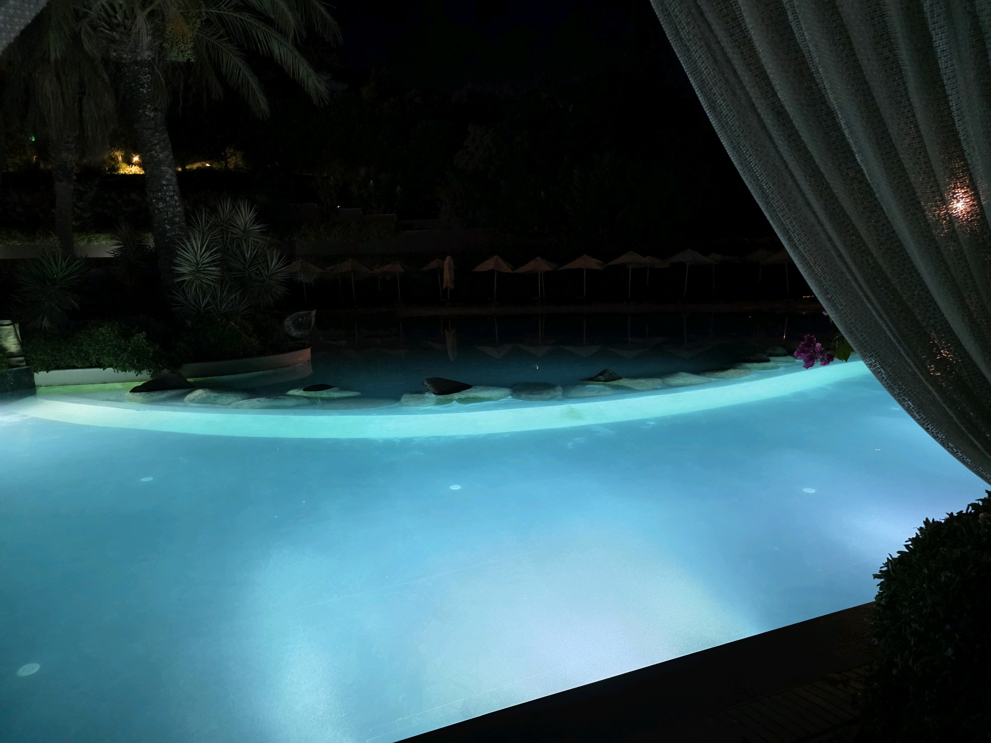 In Bodrum, Turkey, at night, a swimming pool is illuminated by underwater lights, casting a vibrant blue glow. The pool is situated amidst lush greenery, with palm trees and various plants forming a natural backdrop. In the distance, a row of empty lounge chairs with umbrellas suggests a daytime setting that has transitioned into evening. A light-colored curtain partially obscures the view on the right side of the frame, adding a sense of enclosure and intimacy to the scene. The water is calm and reflects the ambient light, creating a serene and inviting atmosphere.