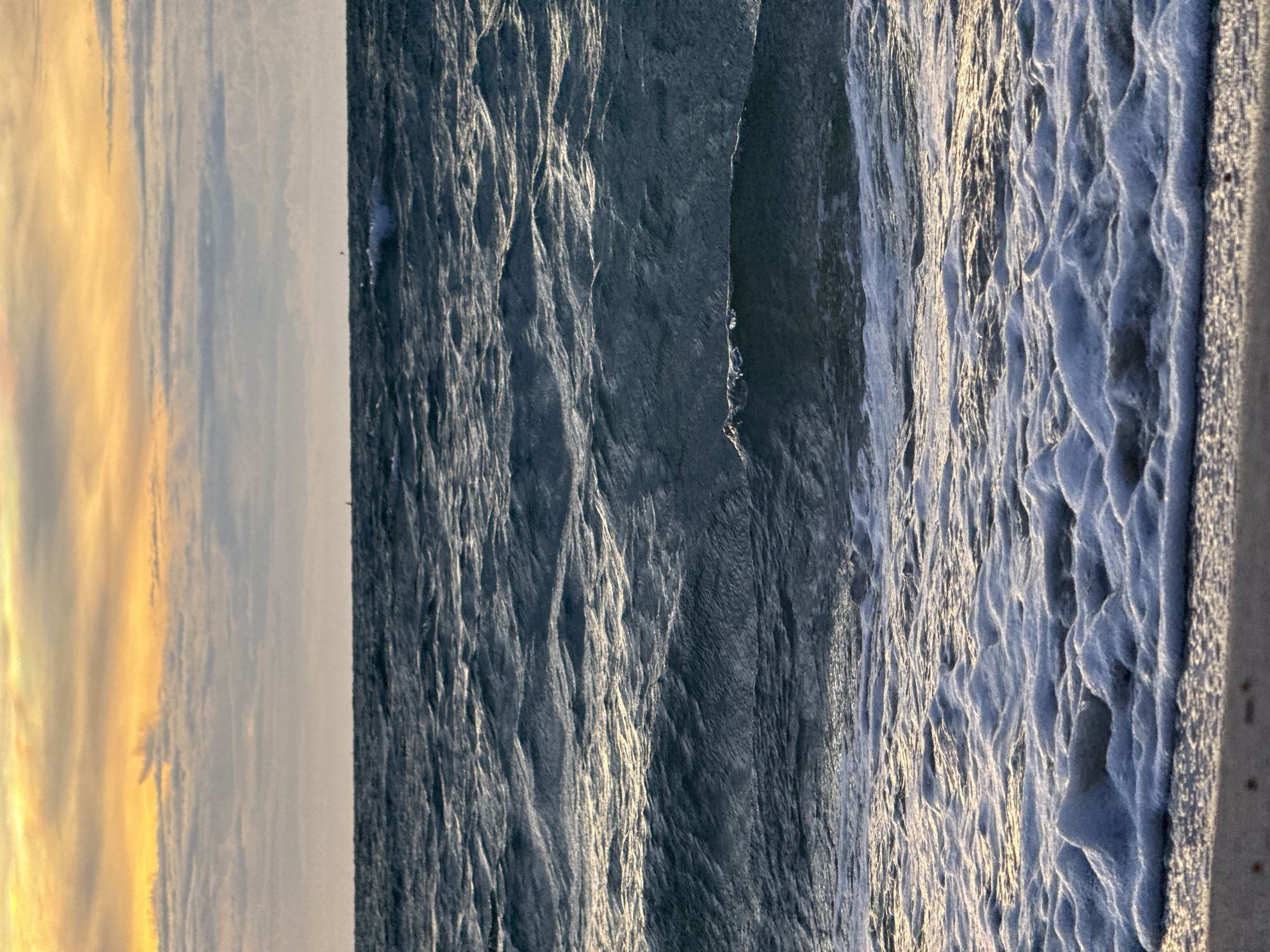 The image shows a coastal scene, likely a beach in Baan Tai, Thailand. The left side features a sunset sky with yellow and grey hues. The middle ground is dominated by ocean waves with reflective light on the surface, transitioning from dark to foamy white as they approach the shore. The extreme right shows a portion of the beach sand. No people are visible.