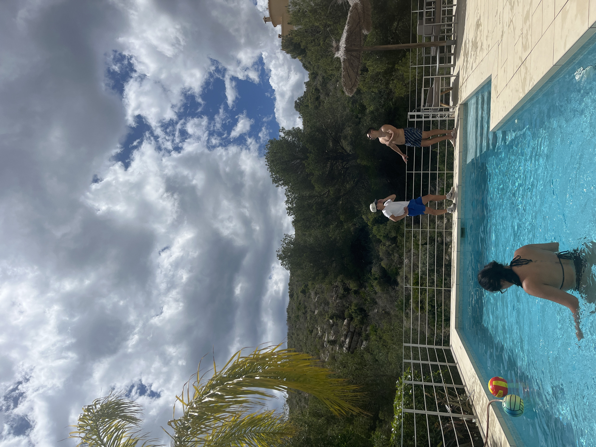 This outdoor scene shows a swimming pool located in el Poble Nou de Benitatxell / Benitachell, Spain. Three individuals are visible. A woman swims in the pool, while two men stand at the edge. A white metal railing borders the pool. Palm trees, foliage, and a hill are in the background, along with a cloudy sky.