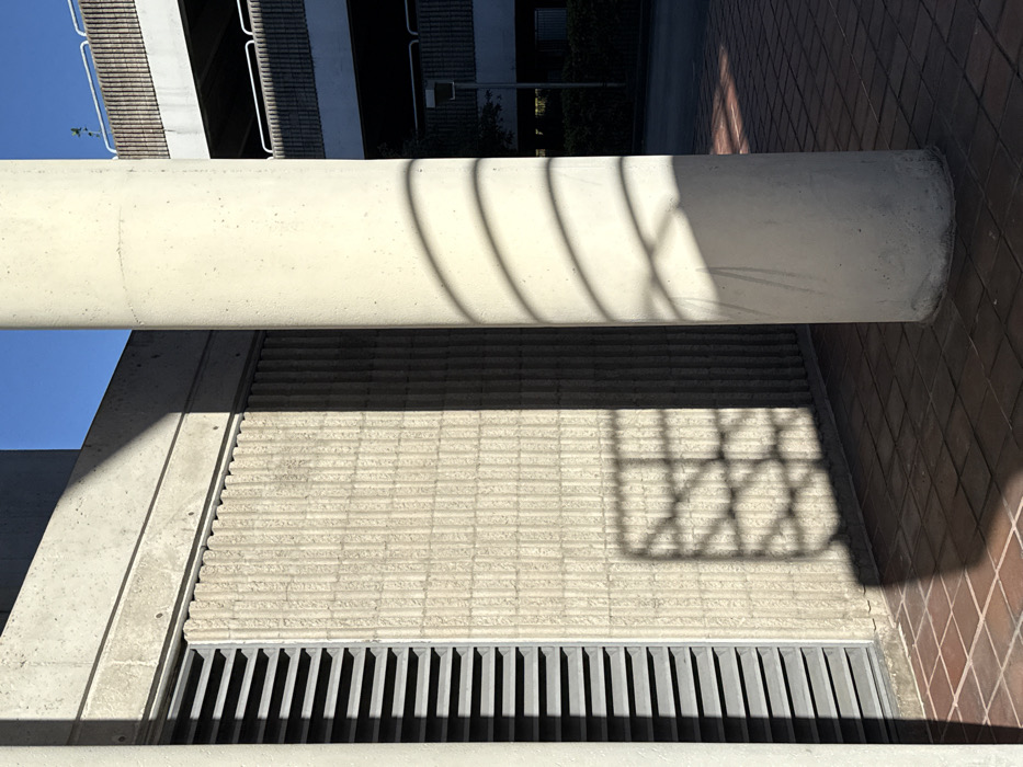 An abstract composition features a cylindrical concrete column horizontally aligned, casting curved shadows upon a textured wall beneath. Below, a horizontal grating with parallel black slats defines a lower plane. A complex geometric shadow resembling a window frame overlaps the textured wall, which transitions into a brick pattern on the right side. The entire scene is bathed in bright light, highlighting the interplay of shadows and textures.
