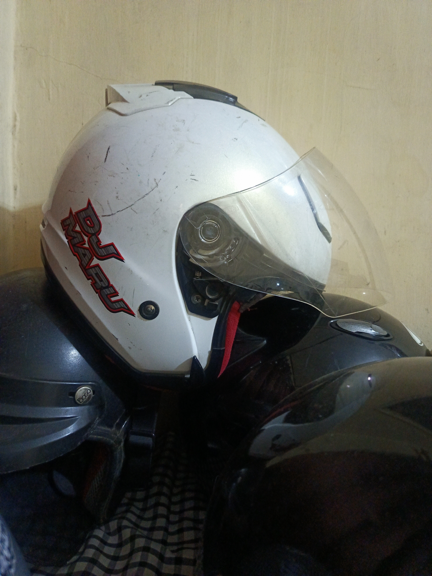 This image shows a pile of motorcycle helmets. The top helmet is white with red and black lettering that appears to say "DJ MARU." It has a clear visor. Underneath the white helmet are at least two other helmets, one dark gray and one black. The helmets are stacked on what appears to be a checkered cloth. The background is a beige, textured wall. The scene is an indoor environment, likely a storage area or a home in the city of Margajaya, Indonesia.