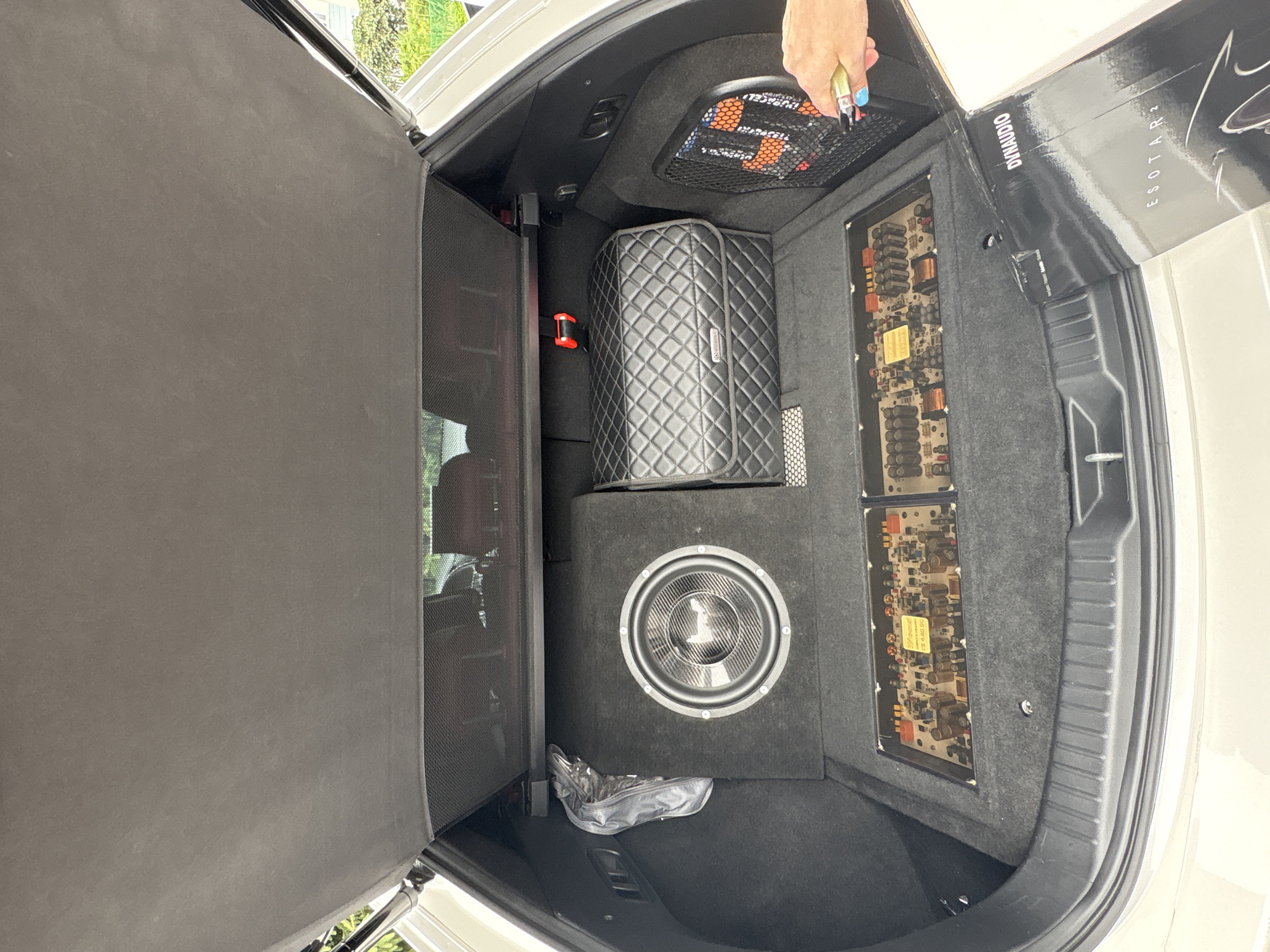The image shows the open trunk of a white car, likely a station wagon or SUV. The trunk is fitted with a custom car audio system. In the left rear of the trunk is a black subwoofer enclosure with a visible woofer. To the right of the subwoofer is a diamond-stitched black quilted storage box. In the foreground, laid flat on the trunk floor, are two electronic components with exposed circuit boards, appearing to be amplifiers. A black box with "DYNAUDIO ESOTAR 2" printed on it is partially visible in the lower right corner, being held by a person whose hand and arm are in the frame. The background shows the car's interior and the trunk lid. The lighting suggests an outdoor setting, possibly during the day. The scene is clean and organized, indicating a focus on the audio equipment. There are no obvious signs of the location being Zanevka, Russia.