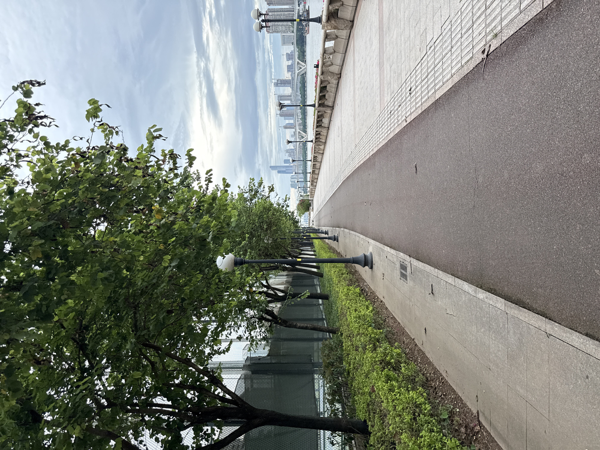 The image depicts a paved pedestrian pathway running alongside a lush green, manicured hedge and a row of trees. The pathway is divided into two sections: a darker asphalt surface on the left and a lighter, tiled surface on the right. Numerous lampposts are spaced along the tiled path. Beyond the tiled path, a decorative stone embankment overlooks a wide river. In the distance, across the river, a cityscape is visible, featuring numerous tall buildings and a bridge spanning the water. The sky is overcast with clouds. The scene appears to be a waterfront promenade in Tianhe District, China, likely a public park or recreational area designed for walking and enjoying the urban riverfront. No people are visible in the immediate vicinity of the pathway.