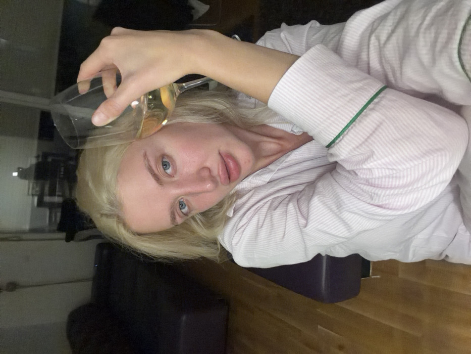 The image depicts a young adult female with blonde hair and blue eyes, looking directly at the camera. She is wearing a light pink and white striped long-sleeved shirt with green piping on the cuffs. Her right hand holds a wine glass containing a pale yellow liquid, consistent with white wine, positioned to the right of her face. The scene is indoors, characterized by dimly lit conditions. On the left, a dark-colored couch is visible, and the floor is composed of light-toned hardwood. In the background, a large reflective surface or window shows a blurred reflection of an interior space containing a table with chairs and a prominent light source. The individual is situated in the city of Budva, Montenegro.