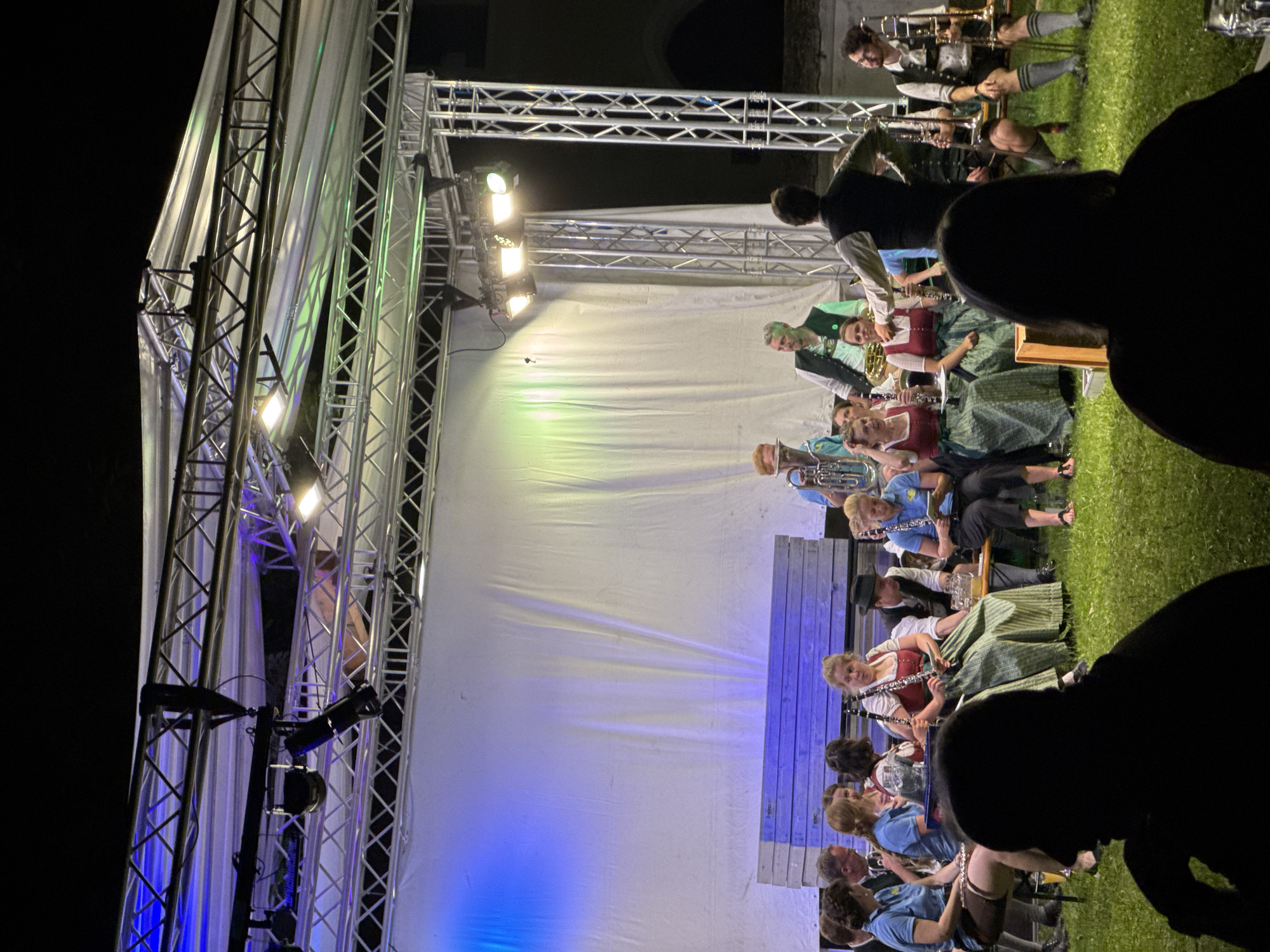 The image depicts an outdoor evening event in Höhenkirchen-Siegertsbrunn, Germany, likely a musical performance. A group of musicians, dressed in traditional Bavarian attire, are seated on a bench on a grassy area. They are playing various instruments, including clarinets and brass instruments. In the background, a stage structure with lighting is visible, with a white backdrop. Some audience members are seated on the grass in the foreground, silhouetted against the stage lights. The overall atmosphere suggests a community gathering or festival.