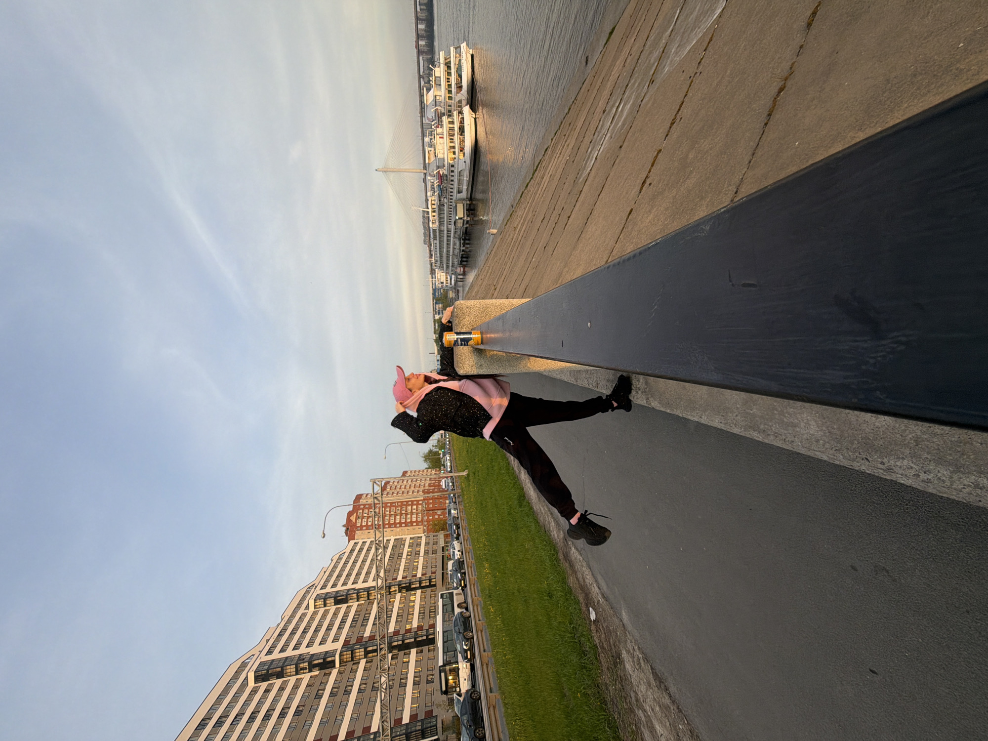 A daytime shot, rotated counterclockwise, shows a person in black pants, black shoes, and a black and pink top, wearing a pink hat, standing on what appears to be a pier or walkway. To their right is a black barrier, then a gray concrete walkway. A strip of green grass is between the walkway and a street with parked cars. Buildings rise on the left side of the image. In the distance, on the right, is water with docked boats and a bridge. The city is Saint Petersburg, Russia.