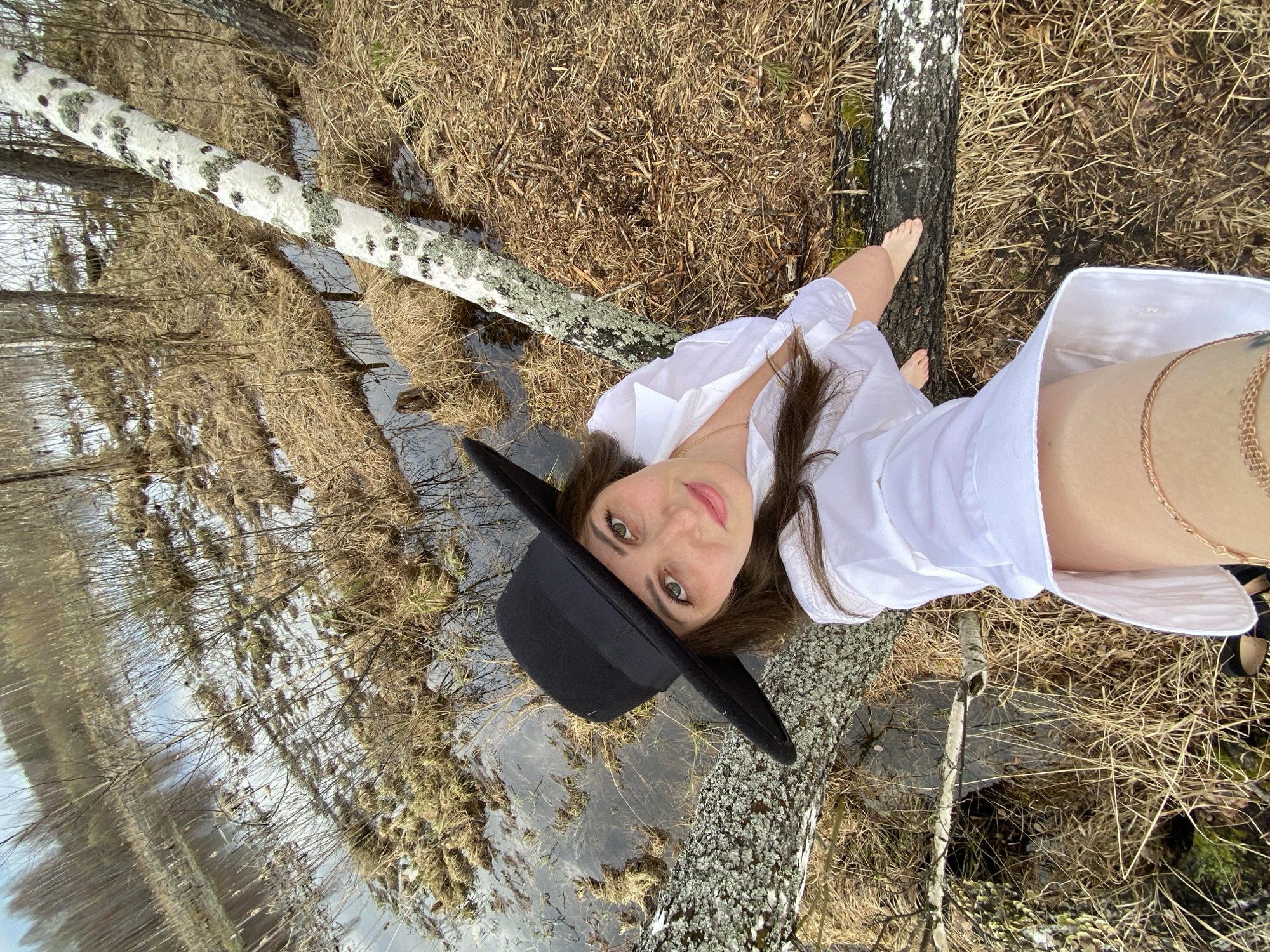 A low-angle, rotated, eye-level photograph depicts a woman in a black hat and a white button-down shirt posing outdoors. The woman has long brown hair, fair skin, and light eyes. She is positioned between two birch trees, one lying horizontally and one standing vertically. The trees are close to a small body of water that reflects the surrounding trees. Dry, brown grass is visible around the water and trees. The photograph was taken near the city of Марковский, Russia.