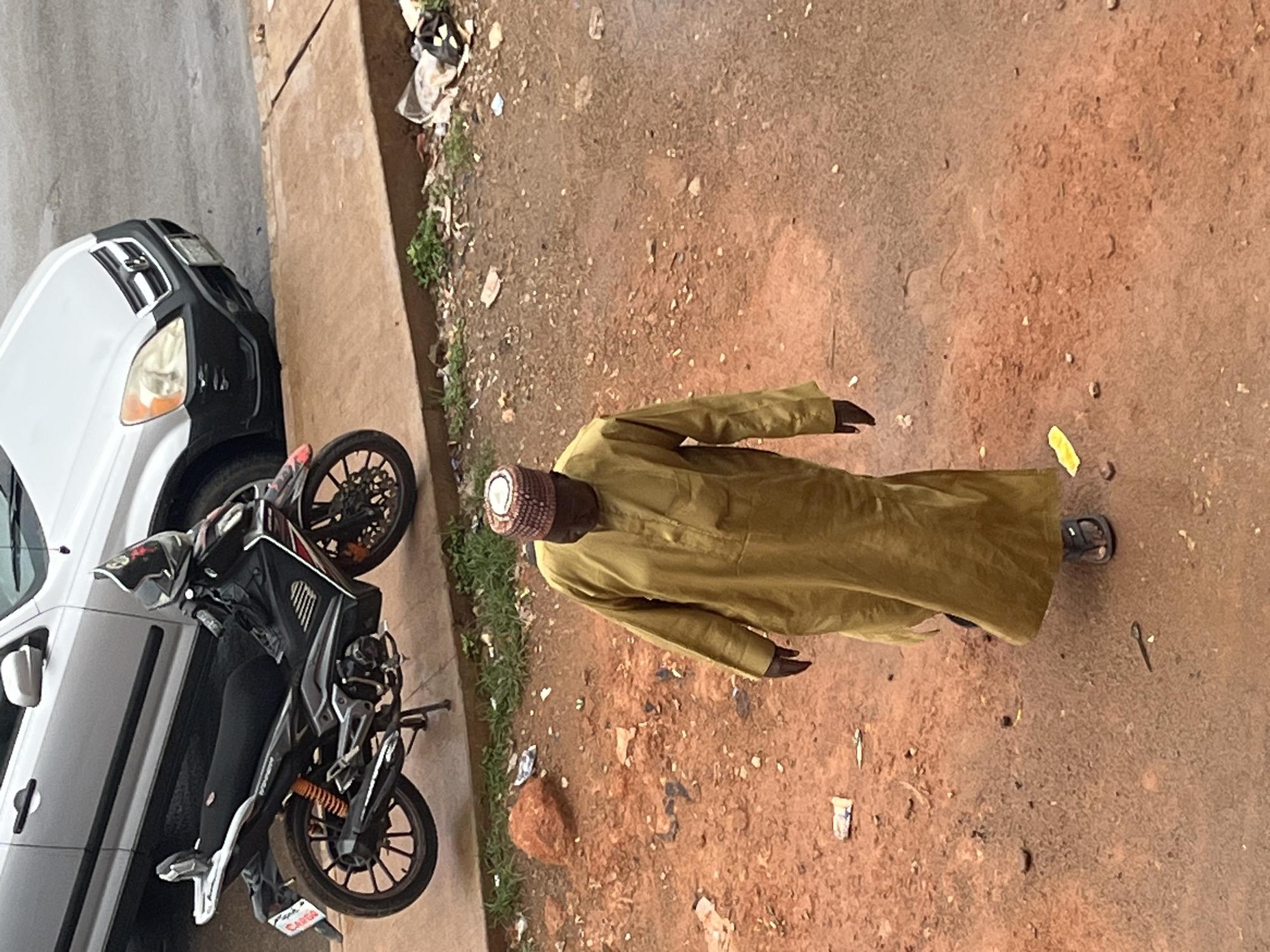 In Jos, Nigeria, an overhead shot captures a man walking on a dirt road. He is wearing a mustard-colored robe and a patterned cap. To his left, a motorcycle is parked on a paved surface, with a silver SUV behind it. The ground is a mix of dirt, pebbles, and scattered debris, including bits of paper and plastic. Green weeds are visible along the edge of the paved area. The scene is illuminated by natural daylight.