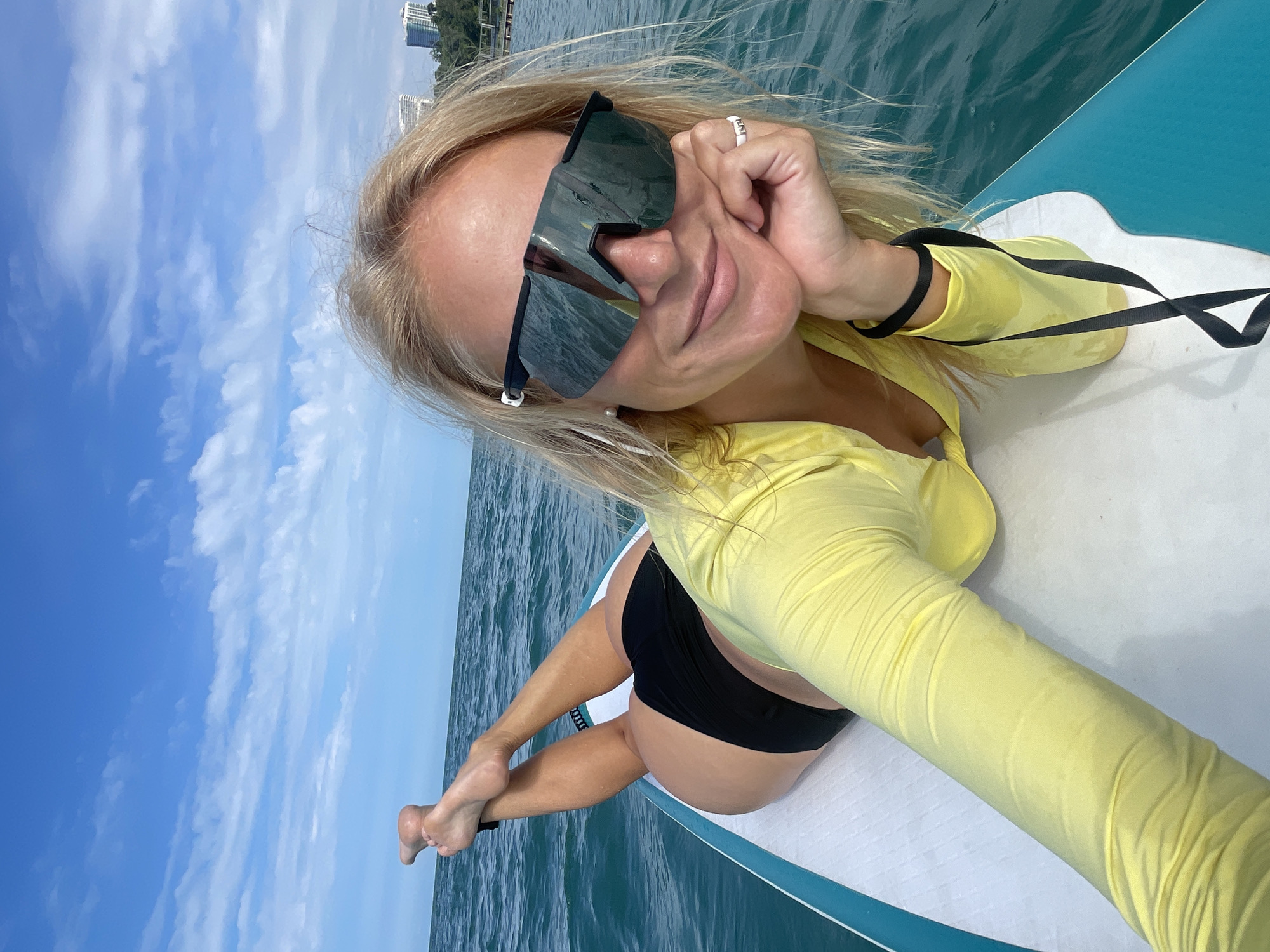 A woman with blonde hair and sunglasses lies on a paddleboard in the sea, with the coastline and buildings of Sochi, Russia visible in the background. The water is blue and calm, with some clouds in the sky. The woman is wearing a yellow rash guard and black bikini bottoms. She is looking at the camera and smiling. Her feet are raised in the air, and her hand is resting on her chin. The paddleboard is white and blue.
