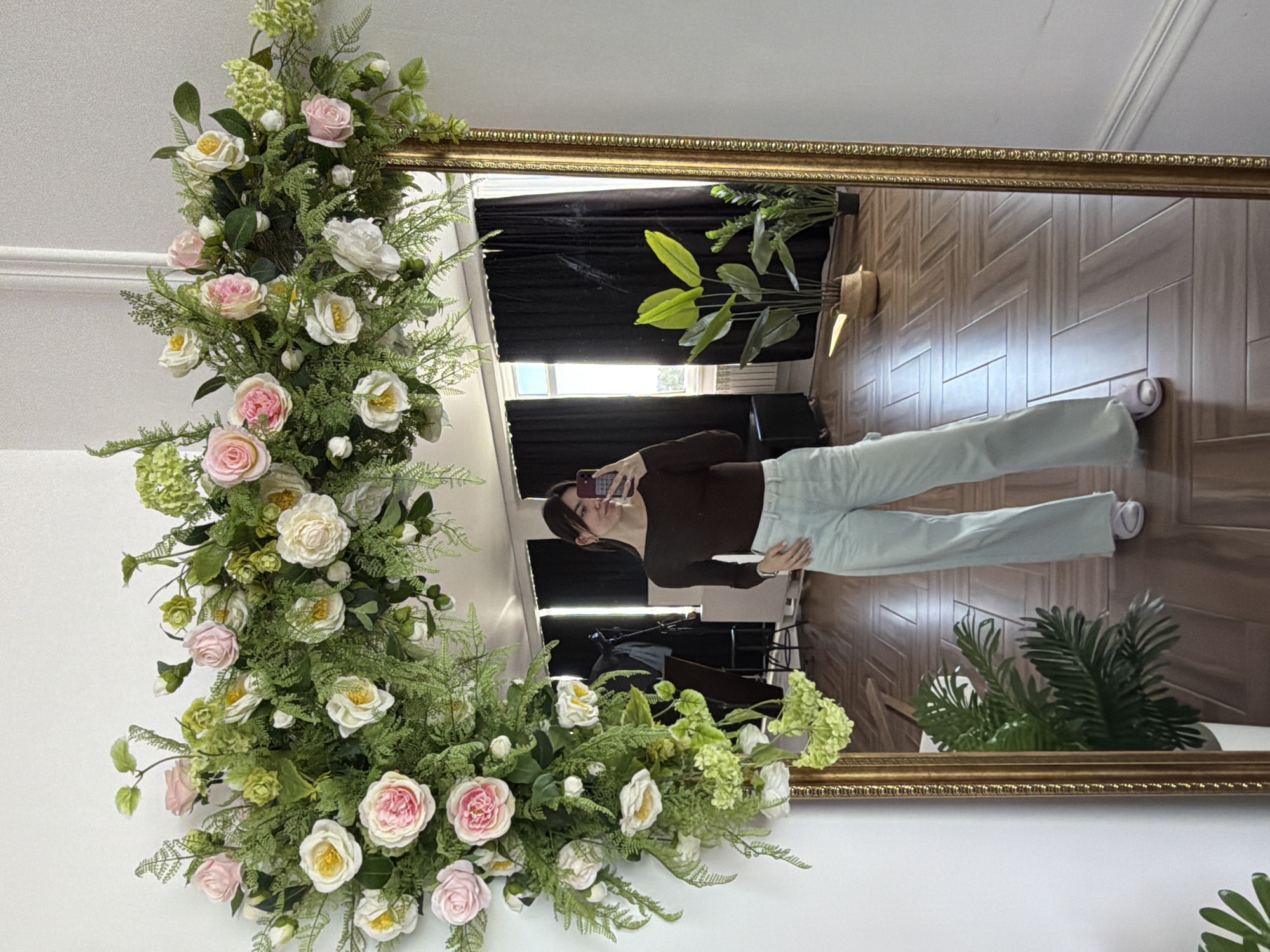 A woman is reflected in an ornate, gold-framed mirror adorned with faux floral arrangements of roses, greenery, and hydrangeas. The reflection reveals a room with hardwood floors laid in a herringbone pattern, a potted plant, and a dark curtain. The woman is holding a cell phone, possibly taking a selfie. She is wearing a brown top and light-colored pants. The background outside the mirror is a plain white wall. The location is likely an indoor space, possibly a residence, in Chaikovsky, Russia.