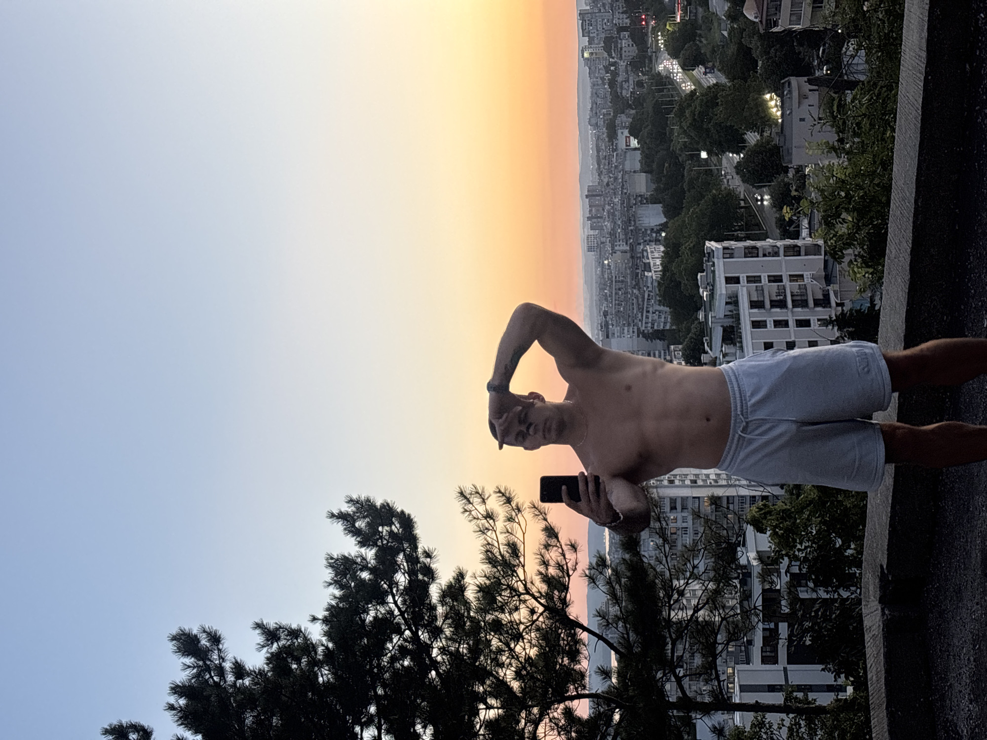 A shirtless man stands on a rooftop in Varna, Bulgaria, holding a smartphone to take a selfie. He is wearing light blue shorts. The man is positioned in the foreground, with a pine tree to his left and a cityscape in the background. The sun is setting, casting a warm orange glow across the sky and illuminating the buildings. The city appears to be on a hill, with cars visible on a road winding through it. The sky transitions from a clear blue at the top to a gradient of orange and pink near the horizon.