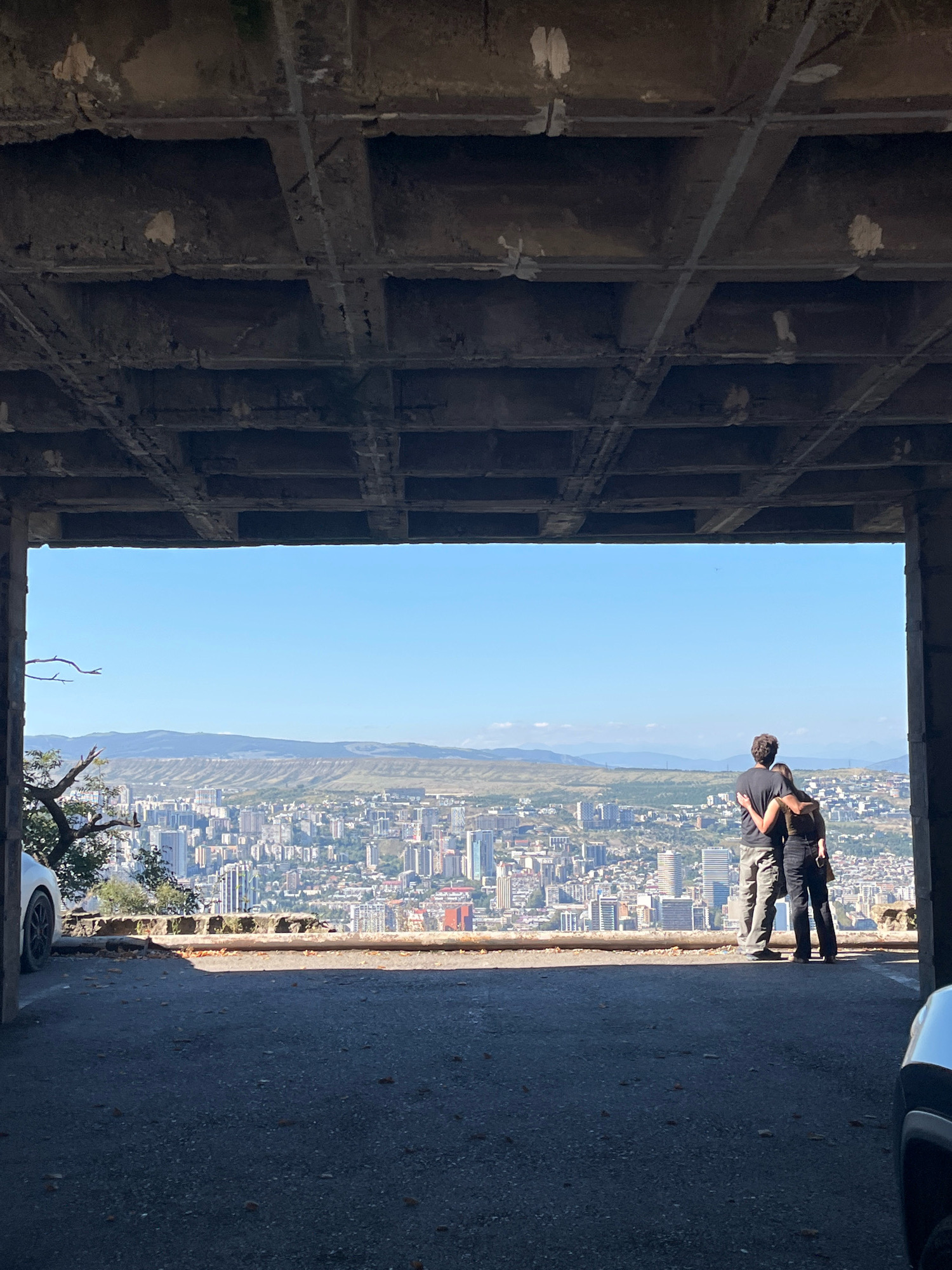 An outdoor, eye-level, medium shot captures a couple embracing and looking out at the cityscape of Tbilisi, Georgia, from an elevated viewpoint. The couple, a man and a woman, stand close together with the man's arm around the woman's shoulders, both facing away from the camera. They are positioned on a paved surface that appears to be a road or parking area, with a concrete structure framing the view. The cityscape below is densely populated with buildings of various heights and architectural styles, stretching out to a hazy horizon with rolling hills. The sky is clear and blue. To the left of the couple, a white car is partially visible. The concrete ceiling of the viewing structure has a grid-like pattern and shows signs of wear and tear. Fallen leaves are scattered on the ground.
