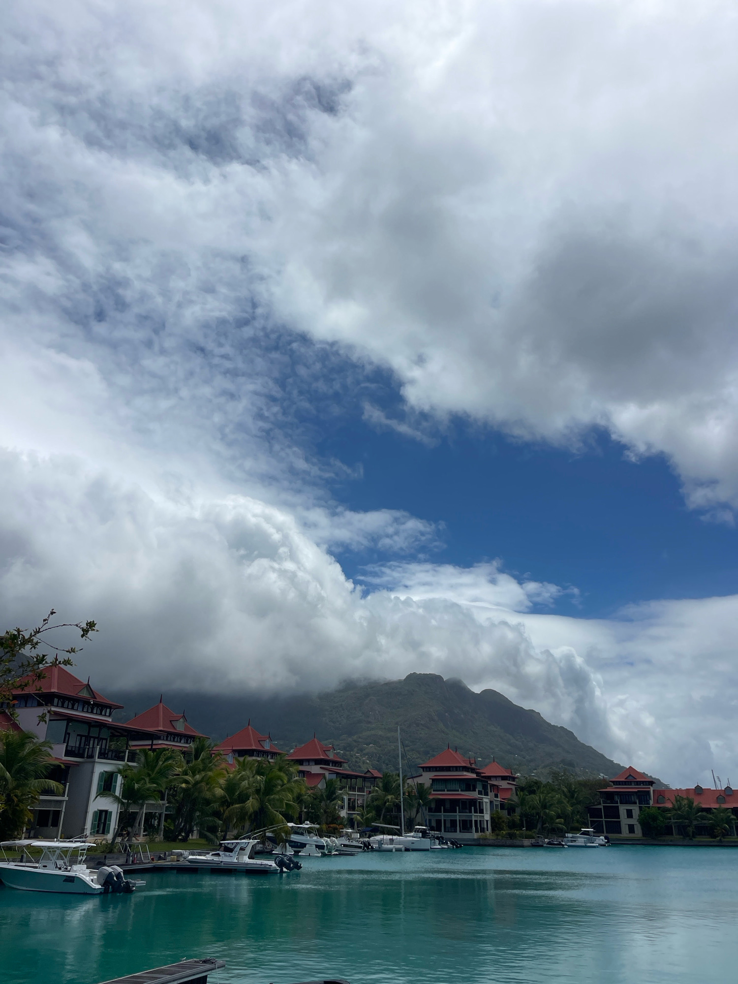 The image captures a serene marina scene in the city of Victoria, Seychelles. In the foreground, a vibrant turquoise body of water is dotted with several white motorboats docked along a wooden pier. The buildings lining the waterfront feature distinctive red-tiled roofs and are interspersed with lush green palm trees, suggesting a tropical resort or residential area. In the background, a majestic mountain range shrouded in dramatic, cloudy skies forms a striking natural backdrop. The interplay of light and shadow across the clouds adds depth and texture to the sky, hinting at the dynamic weather conditions often experienced in this island nation.