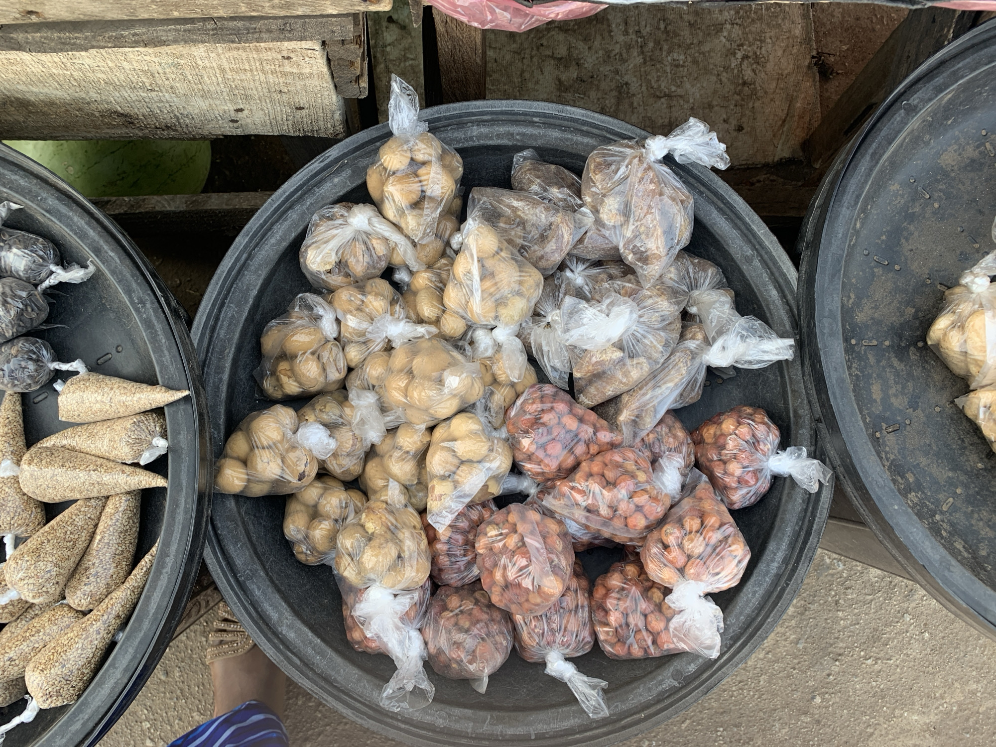 The image shows a top-down view of three large black plastic basins. The central basin is filled with small plastic bags containing various nuts and seeds. The bags in the center basin appear to contain light brown, dark brown, and reddish-brown nuts/seeds. The basin to the left contains cone-shaped bags filled with light tan seeds. The basin to the right contains what looks like white root vegetables in clear bags. The buckets are set on the ground at a produce market near the New Stadium in Nigeria. A wooden shelf is partially visible in the background. A foot with an ankle bracelet wearing blue and white striped clothing is also visible in the bottom left.