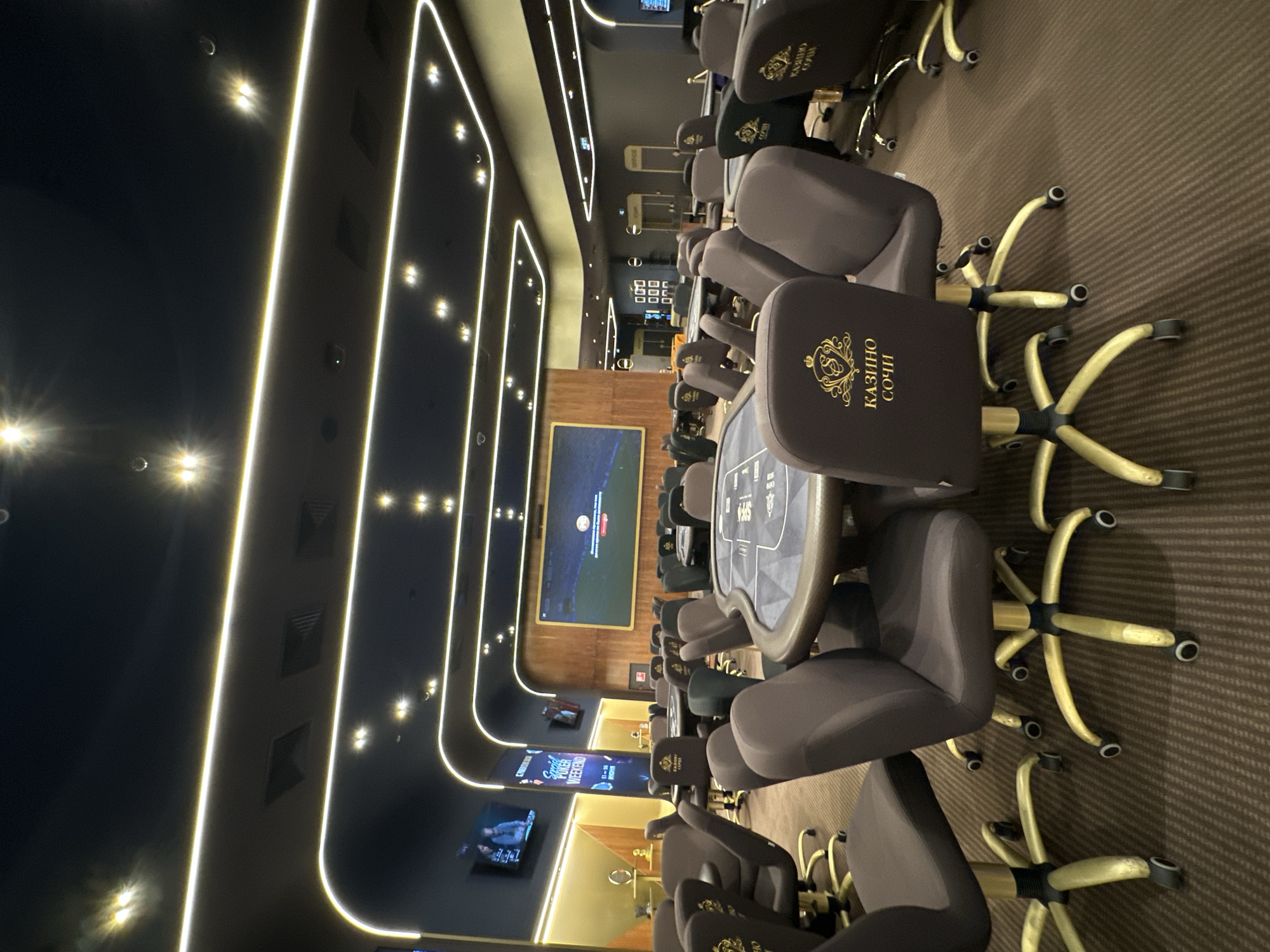 The image depicts the interior of a casino in Estosadok, Russia. The scene is dominated by multiple poker tables, each surrounded by brown upholstered chairs with gold bases and wheels. The tables themselves are round and have a dark blue felt surface with visible markings. In the foreground, one poker table is prominently featured, with several chairs around it. The casino's ceiling is dark and features a modern lighting design with long, curved yellow LED strips that illuminate the space. Several large screens are visible on the walls; one in the background displays a video game or simulation, and another on the left shows a broadcast of two people. The flooring is carpeted in a dark brown, striped pattern. The overall atmosphere suggests a casino designed for poker tournaments or regular play, with a focus on contemporary design and lighting. No people are visible in the image, and no actions or interactions are taking place.