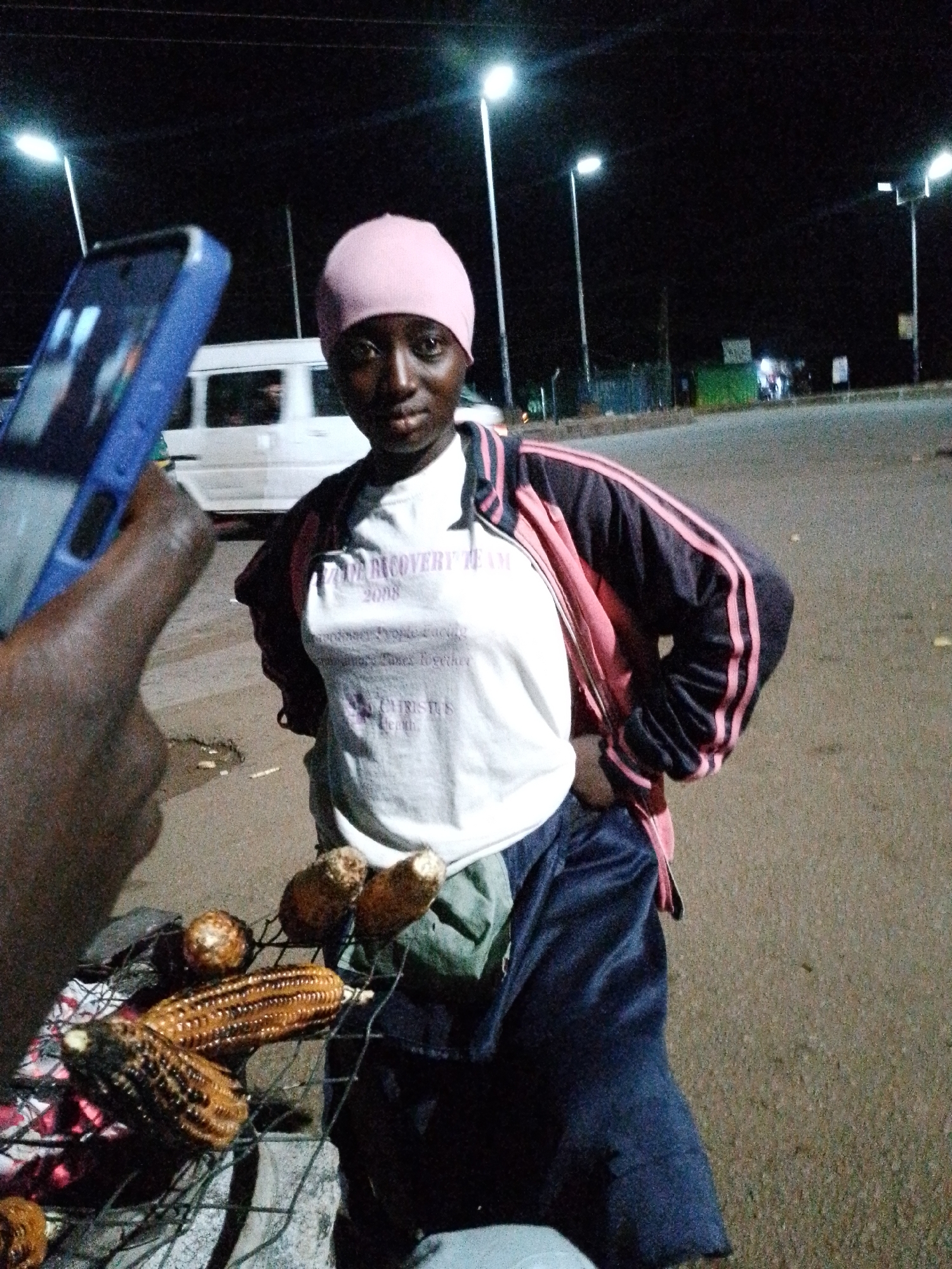 In Bukuru, Nigeria, a young woman stands outdoors at night, holding a phone in her left hand. She is wearing a pink head covering, a white t-shirt with text, and a dark jacket with pink stripes. In front of her, corn is being roasted on a grill over coals. Streetlights illuminate the background, where a road and several buildings are visible. The scene appears to be set on a roadside.
