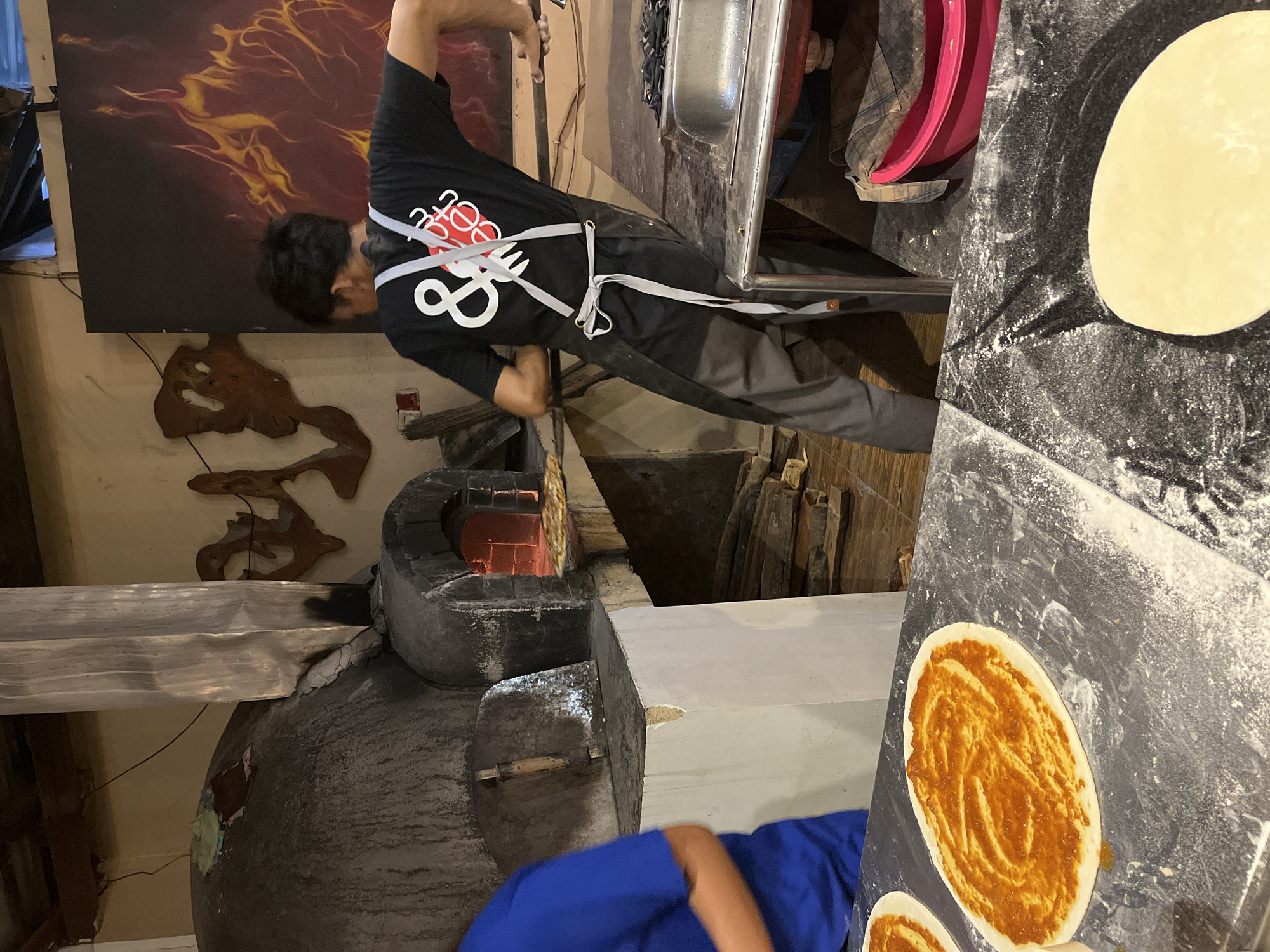 In Karimunjawa, Indonesia, a man wearing a black t-shirt with a white logo and a grey apron is preparing pizzas. He is leaning against a stainless steel counter, his back to the viewer, and his arms are outstretched. One hand is resting on the counter, while the other is near a pizza oven. In front of him, on a floured surface, are two uncooked pizzas, one with a red sauce spread in a spiral pattern, and the other a plain dough disc. The oven, a large, rustic stone structure, is visible in the background with its opening glowing with heat. Inside the oven, a pizza is being cooked. The environment appears to be a rustic kitchen or restaurant setting, with wooden elements and a large painting of flames on the wall. Another person, wearing a blue shirt, is partially visible on the left side of the frame, also near the counter. The scene captures the process of pizza making in a traditional setting.