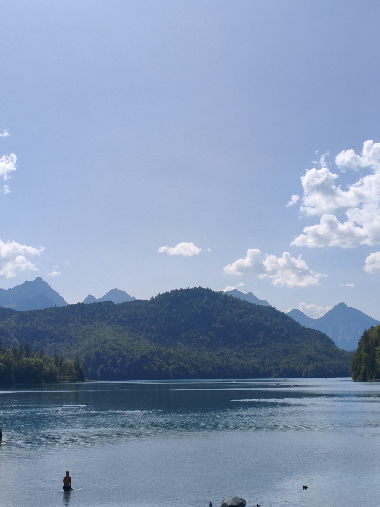 The image depicts a serene lake scene with a backdrop of forested mountains and a bright blue sky dotted with fluffy white clouds. The lake is calm and reflects the sky and surrounding landscape. In the foreground, several people are visible in the water. One person is standing in the water, facing away from the camera, with their torso and head visible. Another person is partially visible on the left edge of the frame, also in the water. Several small boats are scattered across the lake in the middle distance, and some dark shapes, possibly ducks, are swimming on the surface of the water. The overall impression is one of a tranquil summer day in a natural setting. Based on the visual cues of the mountainous landscape and the presence of a large lake, this scene is likely located in the vicinity of Alpsee, a natural lake situated near Hohenschwangau Castle and Neuschwanstein Castle in Schwangau, Germany.