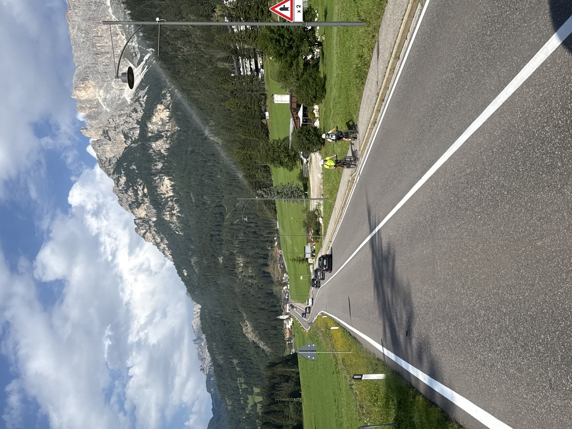 The image depicts a scenic mountain road in Mazzin, Italy, under a partly cloudy sky. A two-lane asphalt road with white lane markings curves through a lush green valley. On the right side of the road, two individuals are cycling on a paved path adjacent to the road. One cyclist wears a bright yellow shirt, and the other wears a dark shirt. Further down the road, several cars are visible, navigating the gentle curves.
The landscape is characterized by steep, forested mountains that rise dramatically in the background. The trees appear dense and dark green, creating a stark contrast with the lighter green of the meadows and fields that flank the road. In the distance, a cluster of buildings, likely part of a village, is nestled in the valley. A prominent church steeple with a white spire is visible among the buildings. A tall, modern street lamp with a curved arm extends over the road on the right side.
To the far right, a red and white triangular traffic sign indicates a sharp curve ahead, with the notation "x 2" suggesting a double curve or increased hazard. The overall atmosphere is one of a serene, picturesque mountain environment, with recreational activity taking place alongside vehicular traffic.