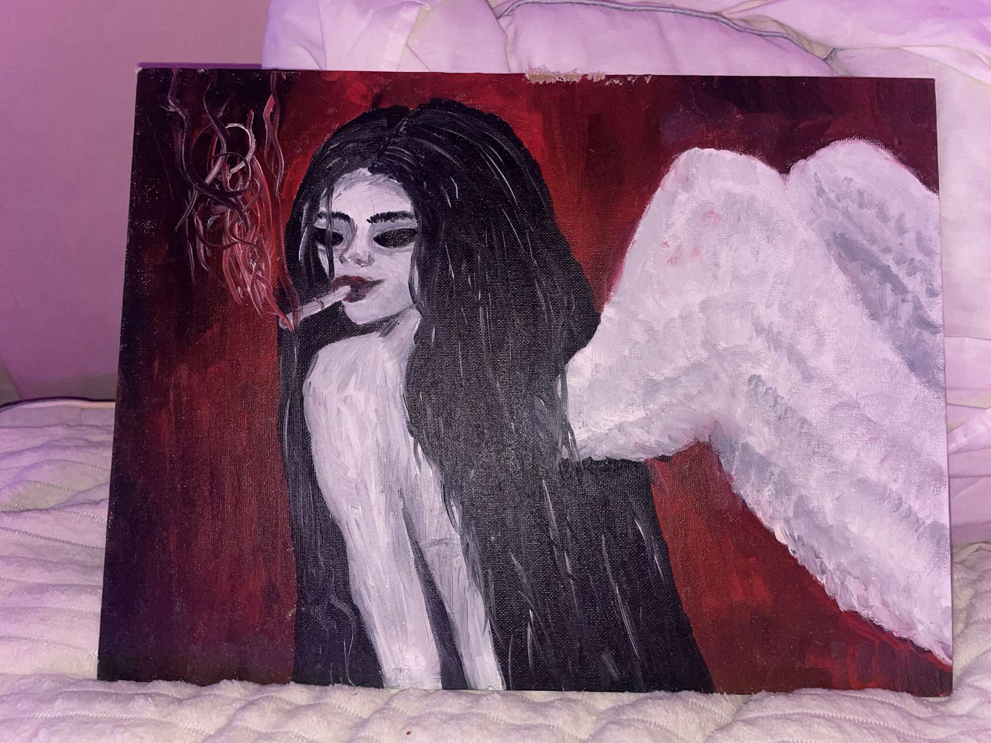This media file features a painting, likely acrylic or oil on canvas, depicting a stylized figure with long, flowing dark hair and pale skin against a dark red background. The central figure, appearing to be a woman, is shown from the chest up, with her upper torso and shoulders exposed. Her eyes are heavily shadowed or painted dark, contrasting with her pale complexion and distinct red lips. She holds a lit cigarette between her lips, and stylized smoke with swirling, organic shapes rises from it, blending into the red background.
To the right of the figure, a prominent set of large, white wings with visible brushstrokes extends across the canvas. The color palette of the painting is predominantly dark red, black, white, and shades of grey. The figure's expression appears melancholic or enigmatic.
The painting itself rests on a white, textured surface, possibly a blanket or duvet, indicating it was photographed indoors. The lighting in the photograph casts a faint pinkish glow on the surrounding bedding. There are no explicit location cues within the painting that suggest Votkinsk, Russia. No visible text is present in the painting or on the canvas.