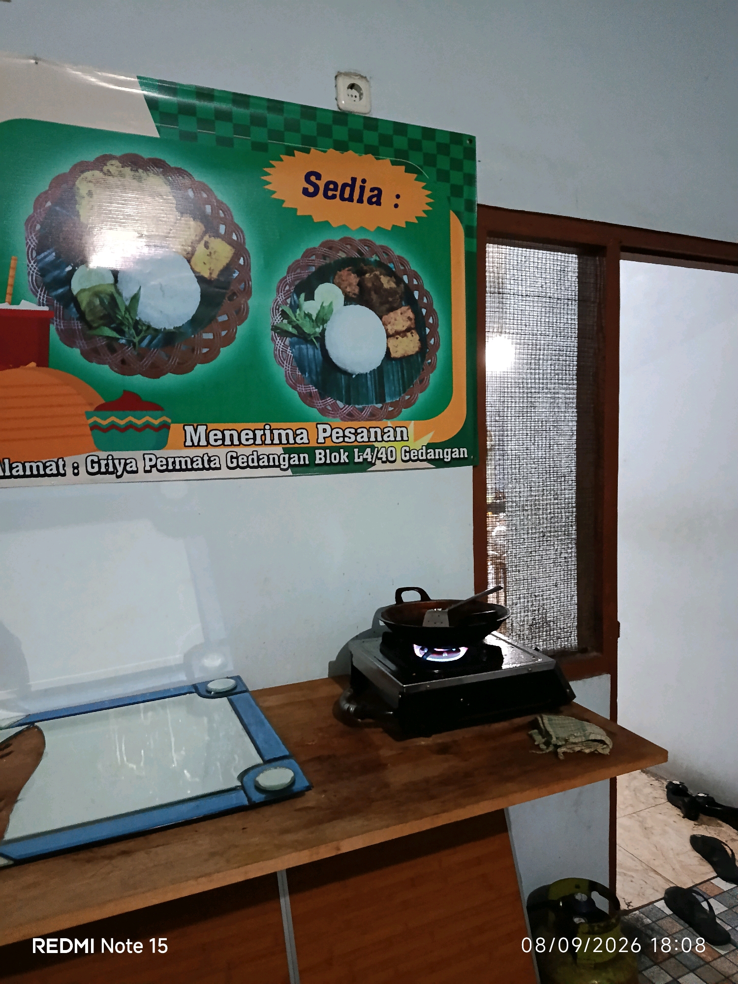 The image depicts a scene inside a modest establishment, possibly a small eatery, in Keboansikep, Indonesia. Dominating the upper portion of the frame is a large, green banner with yellow text and illustrations of food. The banner advertises "Sedia :" (Available:) and "Menerima Pesanan" (Receiving Orders), with details about the address: "Griya Permata Gedangan Blok L4/40 Gedangan."
Below the banner, a wooden countertop is visible. On the countertop sits a portable gas stove with a wok on top, the flame visibly lit. To the left of the stove is a framed mirror with a blue border. Further to the right, a gas cylinder is partially visible, along with a pair of flip-flops on the tiled floor.
The environment appears to be utilitarian, with white walls and a wooden window frame. The lighting suggests it is daytime, with natural light entering through the screened window. No individuals are present in the immediate scene, focusing attention on the setup and promotional material. The overall impression is one of a functional business preparing for or currently serving customers.