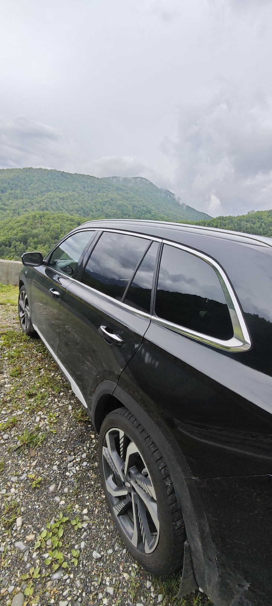 A black car is parked on a gravelly surface near a road, with mountains and a cloudy sky in the background. The mountains are covered in green trees. The location is likely near or in Estosadok, Russia, based on the landscape.
