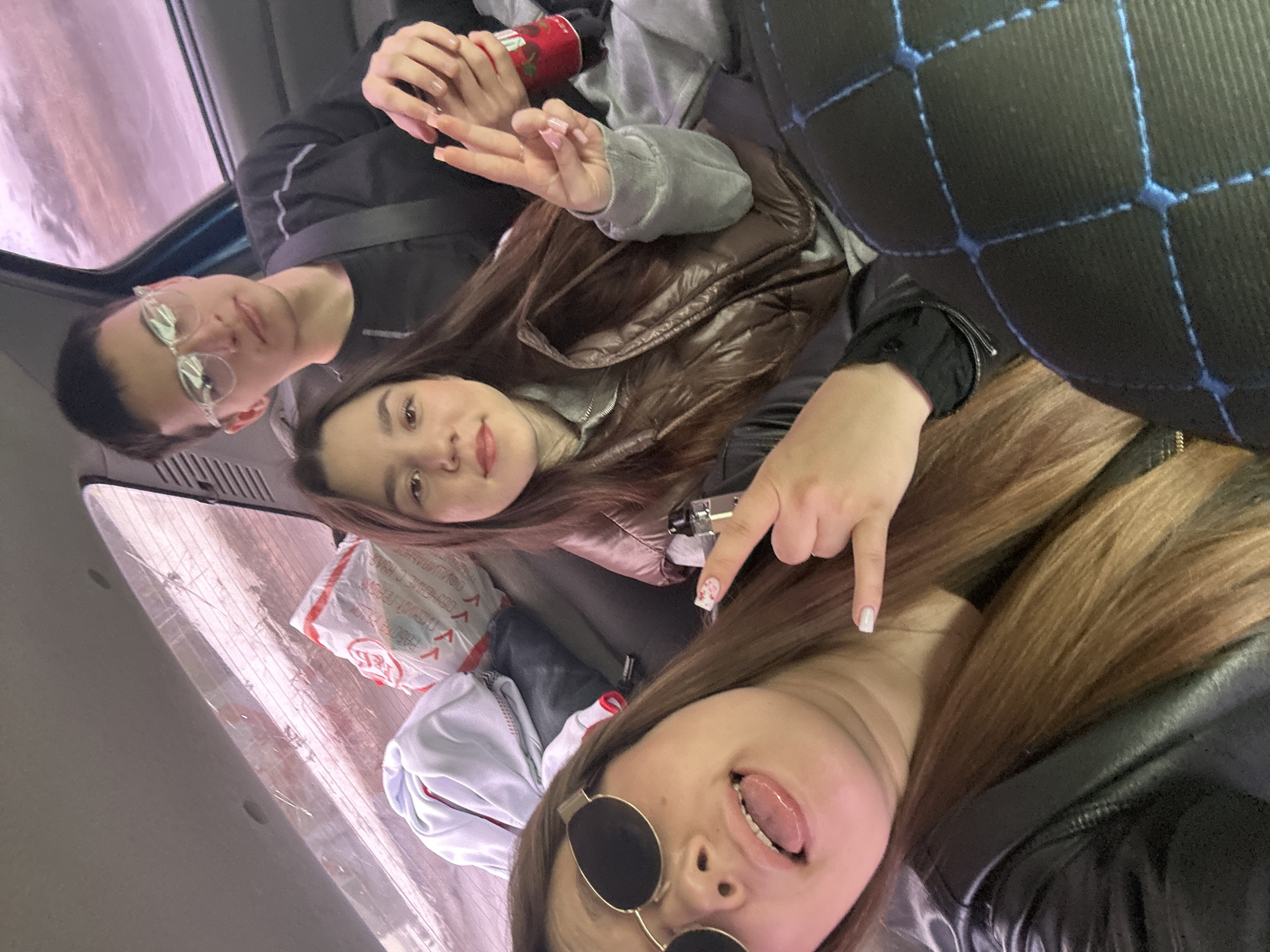 The image shows three young adults posing for a selfie inside a vehicle. The person in the lower portion of the frame is wearing round sunglasses and sticking their tongue out. Above them is another person pointing down with their index finger. The third person, situated towards the top left of the frame, is wearing glasses and looking towards the camera while holding a beverage. Other details include clothing items, a plastic bag with Russian text, and a partial view of the vehicle's interior. The location within the city of Ledukhi, Russia cannot be verified from the image alone.