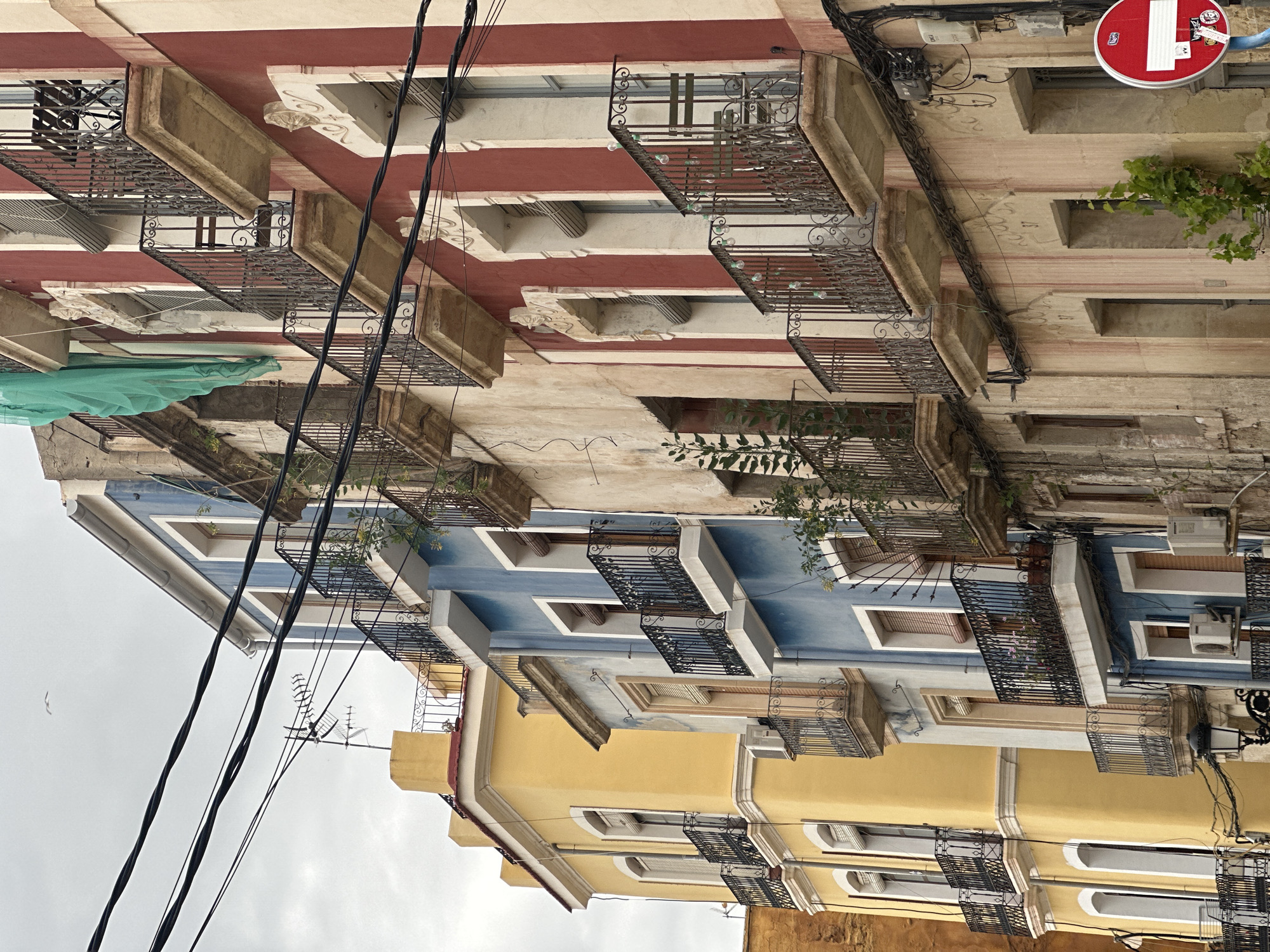 This is an eye-level, medium shot of the facades of buildings. The buildings are a variety of colors, from a red and white striped building to a blue building to a yellow building. All buildings have windows and small wrought iron balconies. A green covering is visible on one of the balconies of the red building. Power lines are strung across the image. A no-entry sign is visible near the top of the image. The location is Unknown, Unknown.
