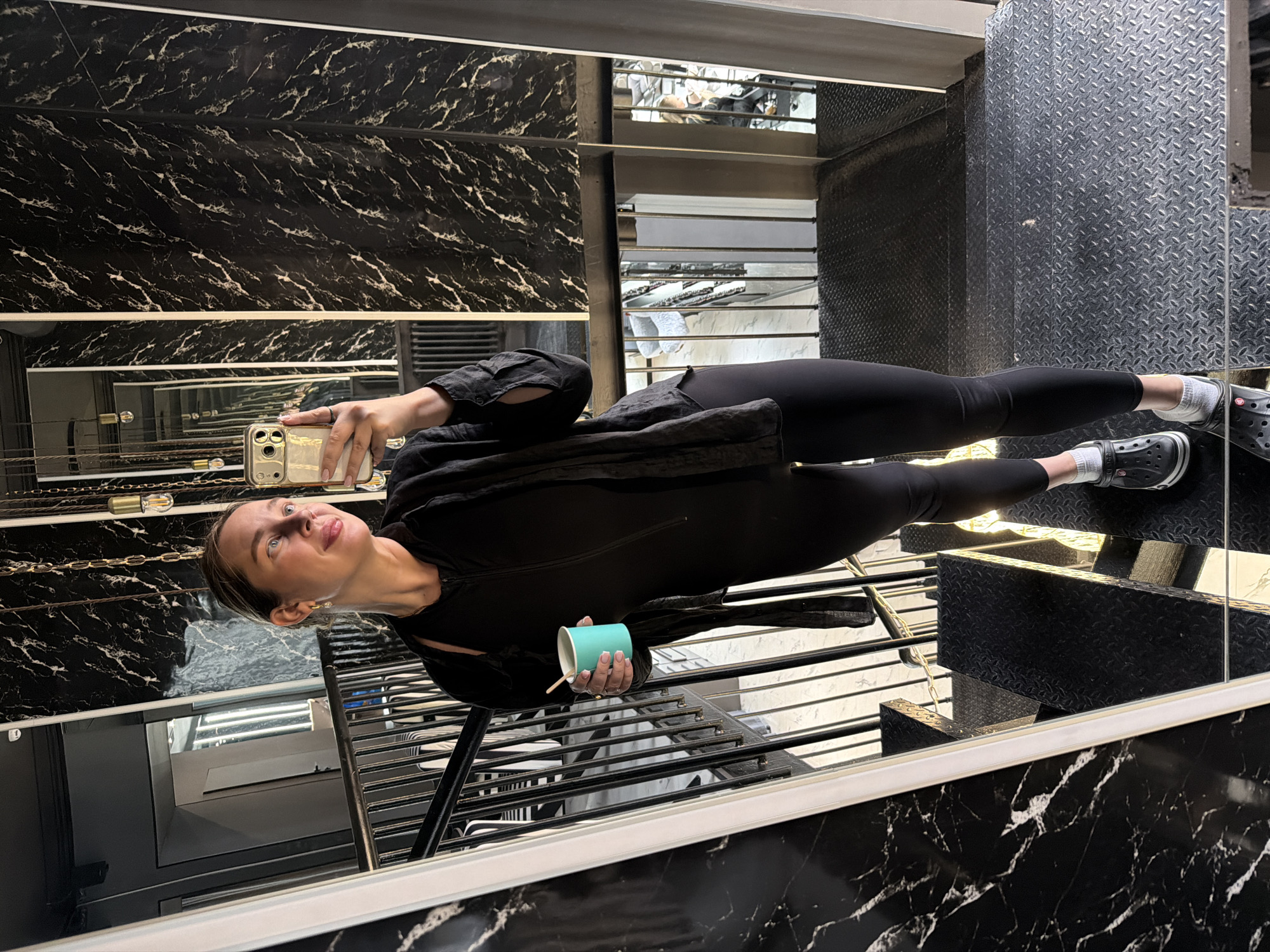 The image shows a reflection of a woman taking a selfie in a mirrored staircase or elevator interior. The walls are decorated with black marble patterned panels separated by white horizontal lines. The staircase is black with a textured metal surface.
The woman is wearing a black outfit consisting of a top, leggings, and black Croc shoes. She is holding a light blue cup in her left hand and her smartphone in her right, positioned to take a selfie. The phone case is gold.
The reflection shows elements of the building's interior, including white walls, railings, and possibly an elevator or staircase above.
Location: Moscow, Russia.