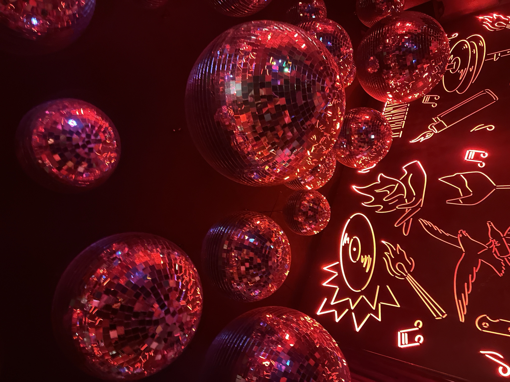 The image shows a dimly lit interior in Milan, Italy. Several disco balls of varying sizes hang against a dark background. To the right of the disco balls, a collection of neon signs, including musical notes, a vinyl record, a lighter, wings, and other artistic doodles, adorns the wall. The entire scene is bathed in a red hue, likely from the neon signs and the reflective surfaces of the disco balls. There are no people visible in the image.