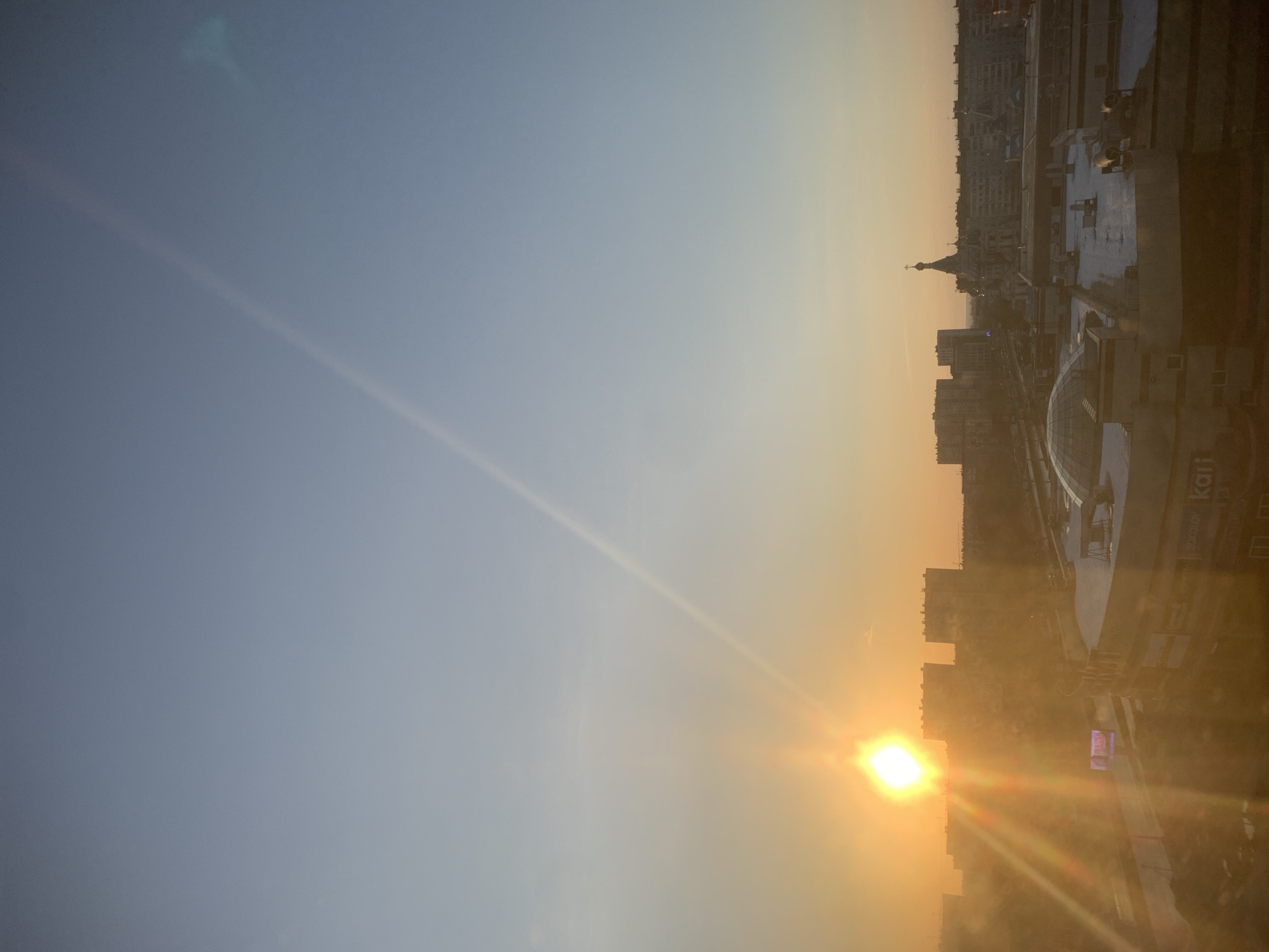 The image shows a cityscape at sunrise or sunset, taken from a high vantage point in Izhevsk, Russia. The sun is positioned low in the sky, casting a warm, golden light across the buildings and the urban landscape. The sky transitions from a light blue at the top to a yellow-orange near the horizon where the sun is located. Buildings and structures are visible in the distance, including a church with a spire, and a modern shopping mall, with a glass dome, on the right side of the frame with "Karl" visible in large letters on its facade.