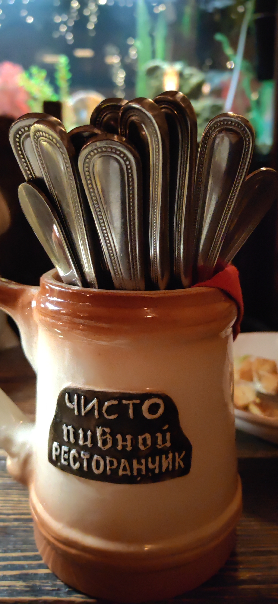 In the foreground, a ceramic stein-like container, possibly a beer mug, holds an assortment of silverware, including spoons and possibly forks, with their handles facing upwards. The container is predominantly white with a brown rim and a decorative brown band around the base. A black label is affixed to the front of the container, featuring white Cyrillic text. The text appears to read "ЧИСТО ПИВНОЙ РЕСТОРАНЧИК", which translates to "CLEAN BEER RESTAURANT". The background is out of focus, showing what appears to be an aquarium with green plants and a light source, suggesting an indoor setting. A blurred plate with food items is visible to the right of the container. The overall scene is a close-up shot focused on the silverware holder. Based on the text on the label, this establishment is likely a beer restaurant located in the city of Homyel, Belarus. No people are visible in the image.