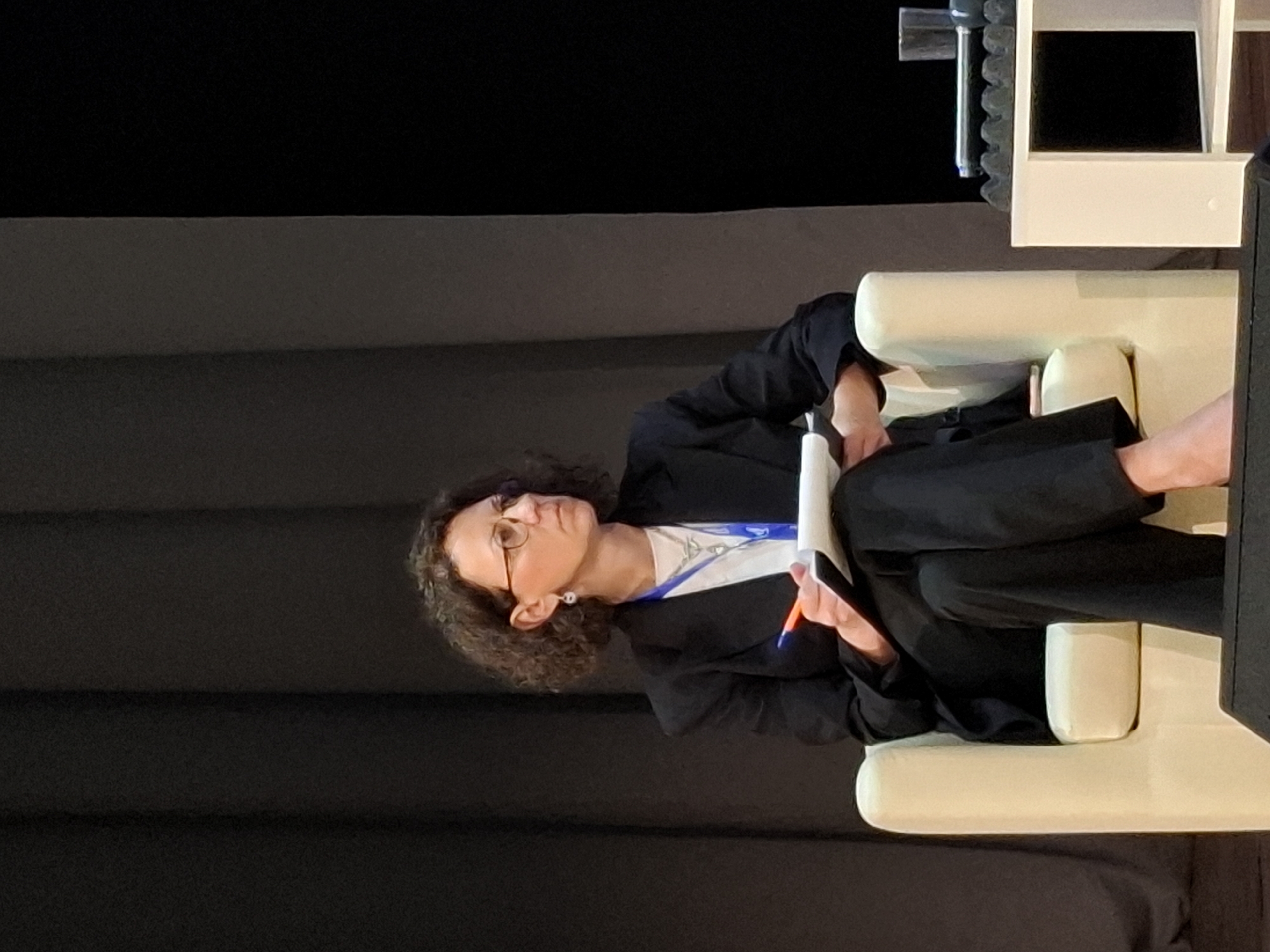 A woman with curly brown hair and glasses sits in a cream-colored armchair on a stage. She is wearing a black suit jacket over a white shirt and black pants. She holds a white notepad and an orange pen in her hands, appearing to be taking notes or writing. She is looking to her right, off-camera. The background is a dark gray curtain. To the right of the armchair is a white table with a glass of water and some electronic equipment on it. The bottom of the image shows a black speaker. The image does not provide any specific information about the location within the city of Kungur, Russia.