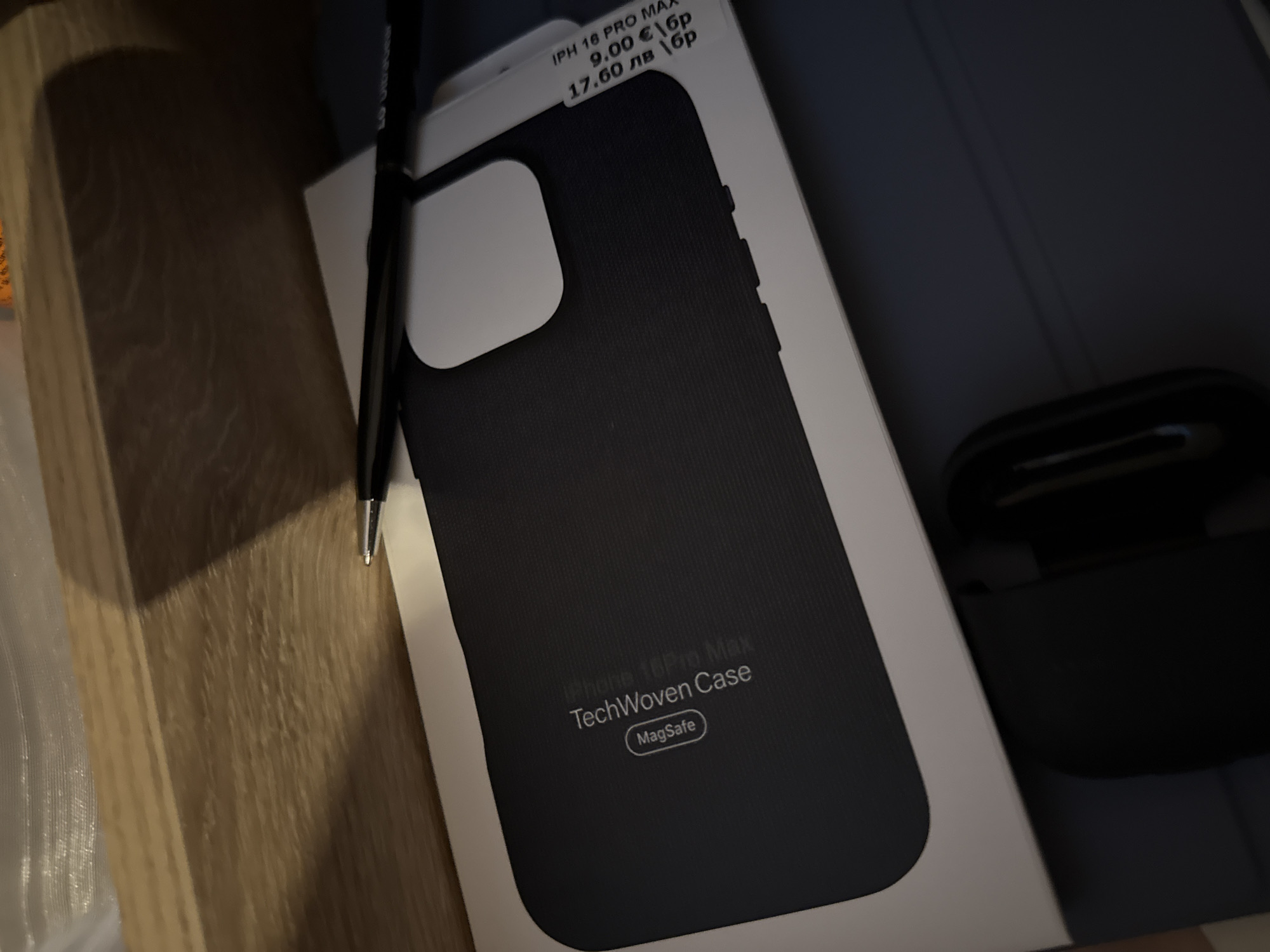 The image shows an "iPhone 16 Pro Max TechWoven Case MagSafe" still in its packaging, which also has a price sticker of 9.00€/ 17.60 лв. A black pen rests across the top of the box. To the right, partially in the frame, is a black charging case for wireless earbuds. The setting appears to be indoors, possibly a home, with a wooden surface visible to the left and a dark blue or grey surface visible in the background. There is no explicit indication of Lovech, Bulgaria.