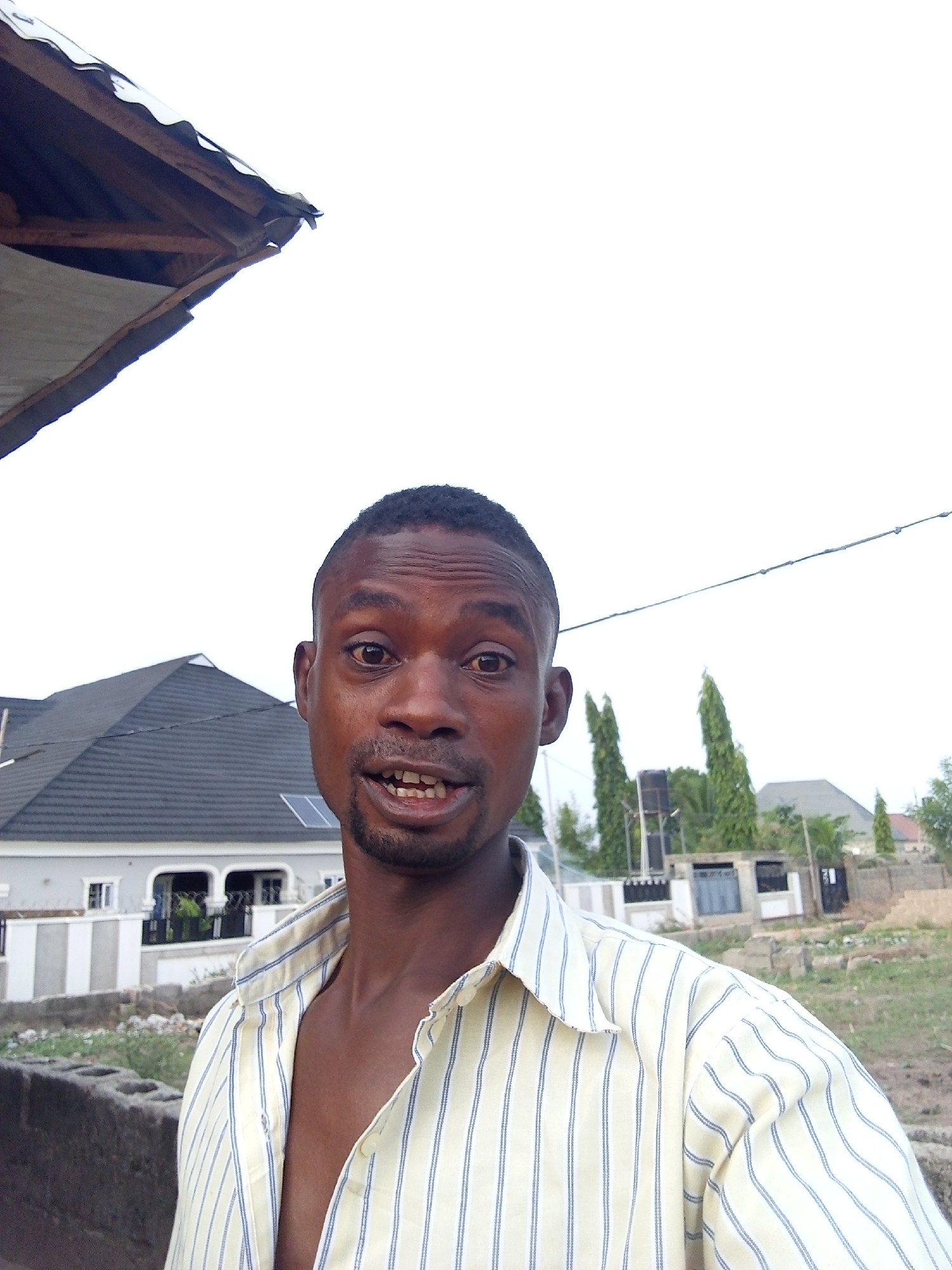 The image shows a man with dark skin and short, cropped hair, wearing a partially unbuttoned, striped shirt with yellow and blue lines. He appears to be standing outdoors in Adonkolo, Nigeria. A single-story house with a dark, steeply pitched roof and a light-colored facade is visible in the background. Trees, a utility pole, and other buildings can also be seen in the distance. An unfinished concrete structure is visible in the foreground. A section of a corrugated metal roof is visible in the upper left corner. The sky is overcast.