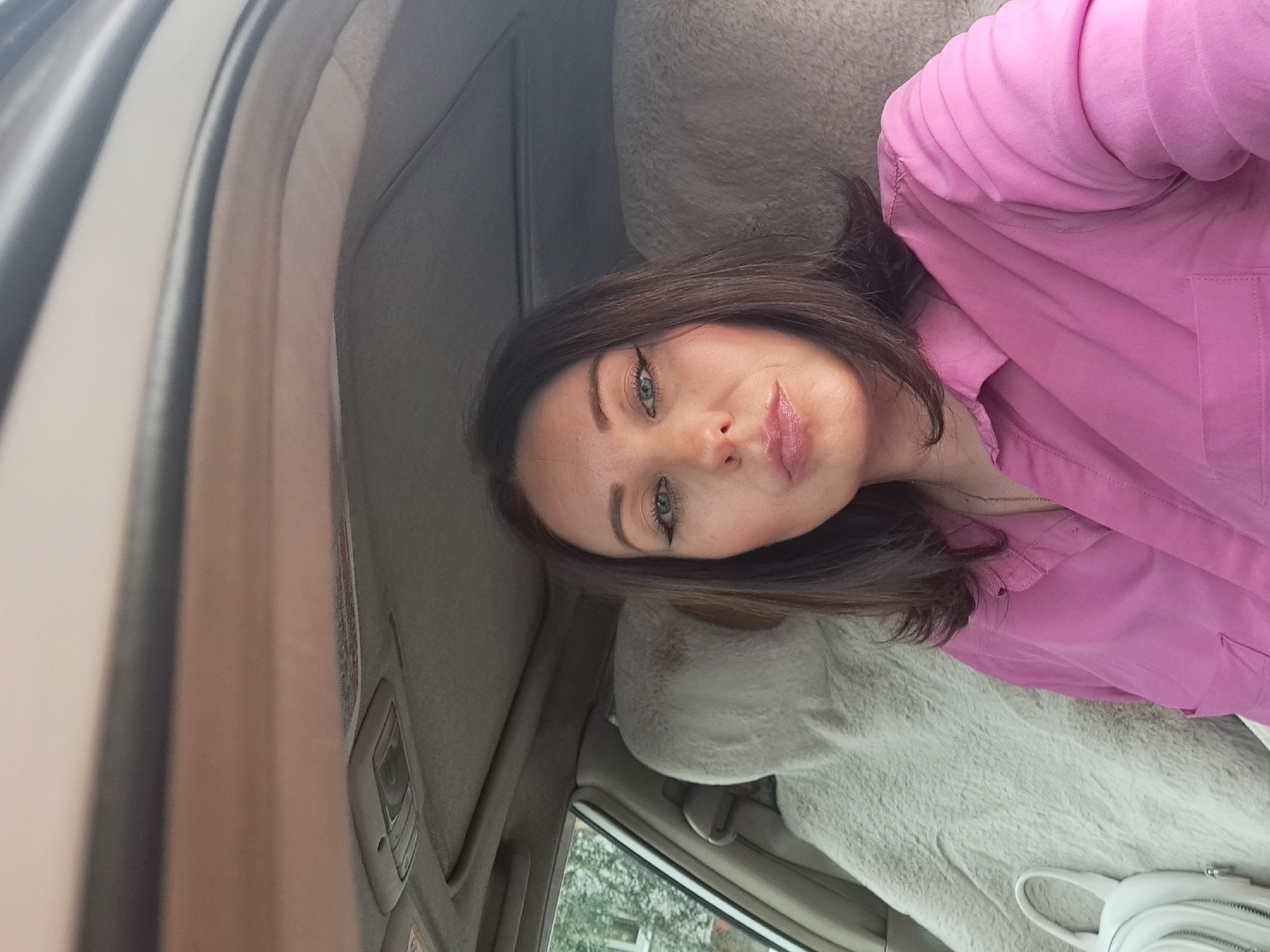 A woman with dark brown hair is sitting in the driver's seat of a car, wearing a pink collared shirt. She is looking at the camera, her face is partially obscured by her hair and the angle of the photo. A white handbag is visible on the passenger seat next to her. The interior of the car is visible, including the ceiling and the headrest of the passenger seat, which is covered with a light-colored sheepskin. The scene is an interior shot of a car, taken from the perspective of the person in the driver's seat. The location is not specified in the image, and therefore, it cannot be determined to be in Irkutsk, Russia.