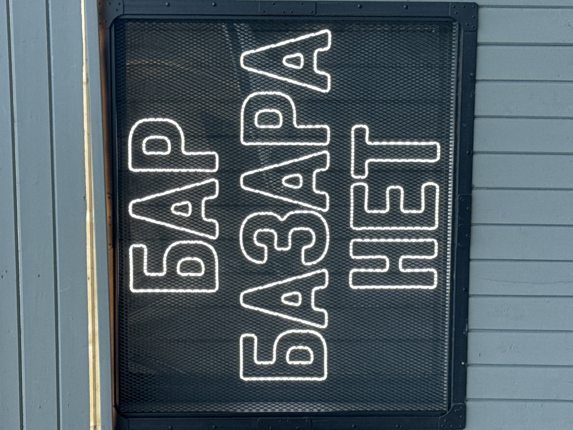 The image shows a neon sign displaying "БАР БАЗАРА НЕТ" in white letters against a dark mesh background. The sign is mounted on a light blue wooden wall. The neon sign translates to "Bar no market" or "No market bar". The sign is framed by black metal and appears to be outdoors. The style of the sign and the wooden wall suggests a casual establishment, possibly a bar or cafe, in an urban setting. The presence of Cyrillic script indicates a location where Russian is spoken, such as Saint Petersburg, Russia. The image is taken from a slightly angled perspective, looking up at the sign.