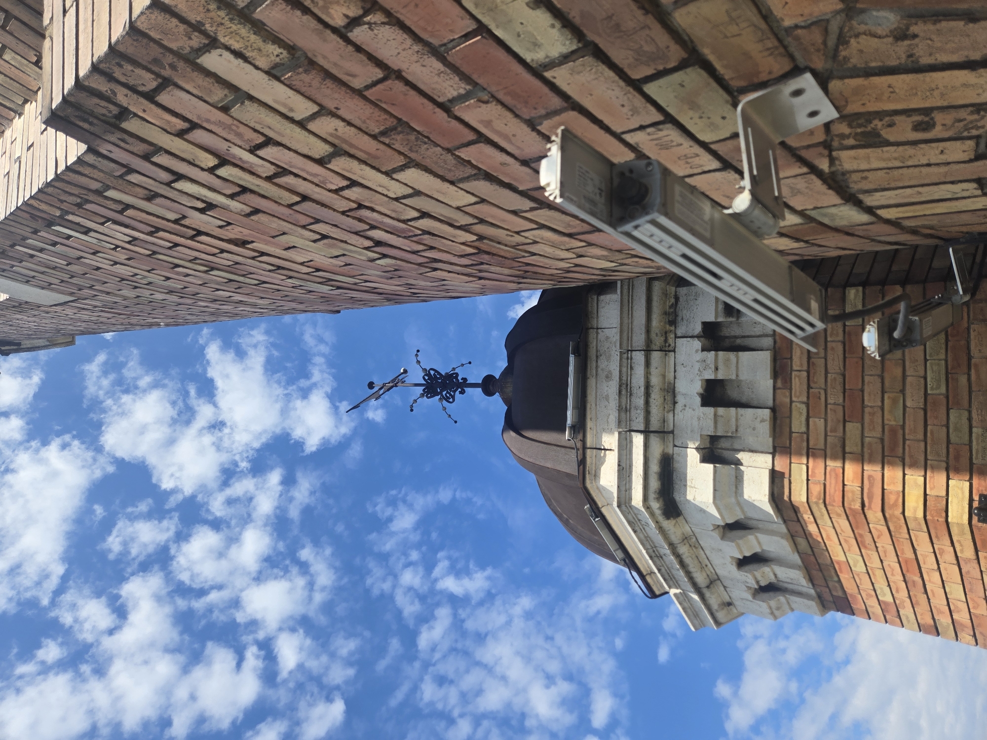 The image, taken from a low angle, shows a view between two buildings in Zemun, Serbia, under a blue sky with scattered clouds. The buildings are constructed of red brick, with the upper building exhibiting an arching facade. Modern lighting fixtures are mounted on the brick structure. Between the buildings, a steeple with an ornate weather vane is visible. The style of architecture suggests a historic urban setting.