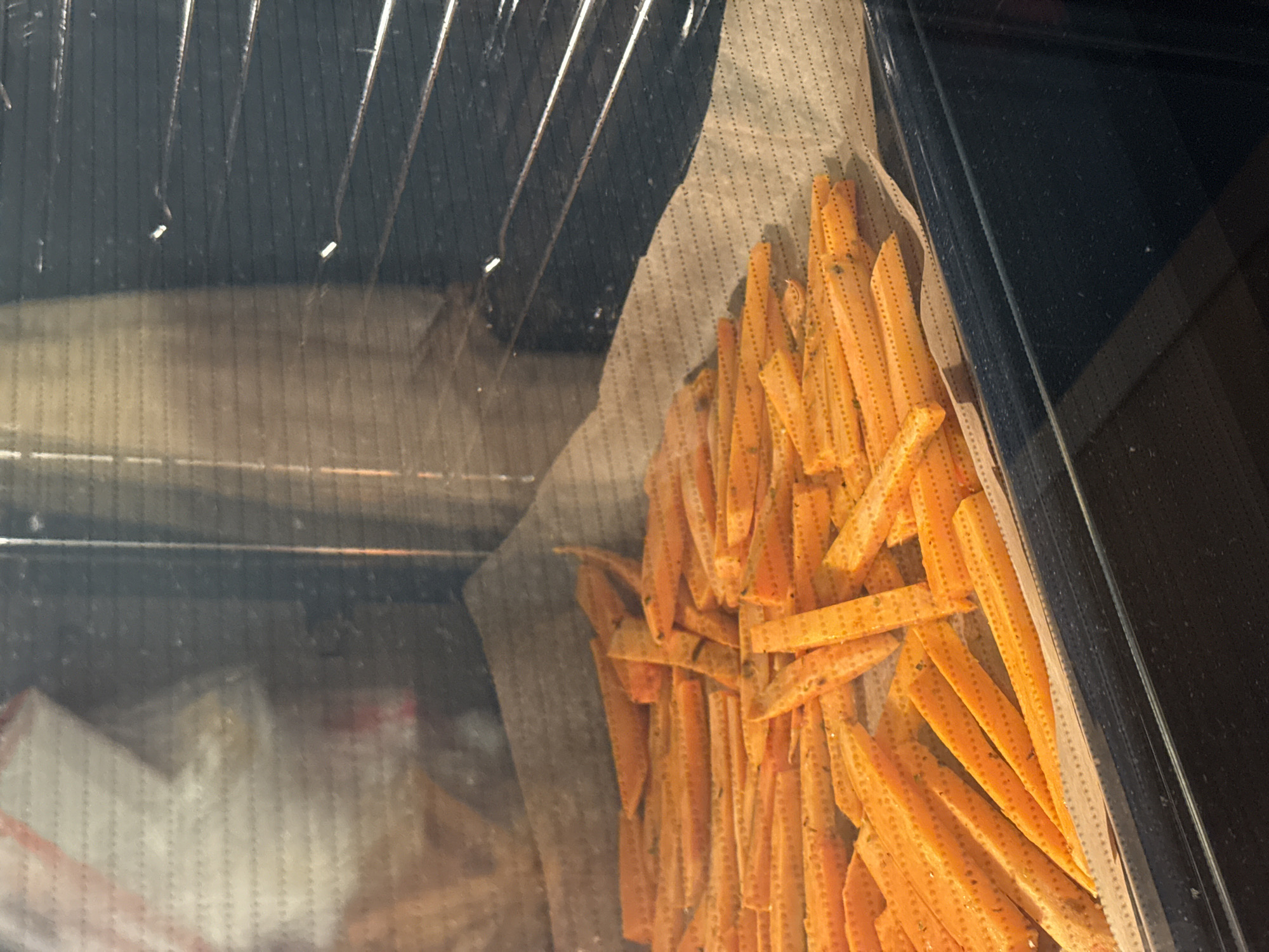 A close-up, interior shot taken through the glass door of an oven. Inside the oven, orange-colored, stick-shaped objects, likely sweet potato fries or a similar food item, are scattered on a sheet of parchment paper. The rack of the oven is visible through the glass. The location or city is not discernible from the provided image.