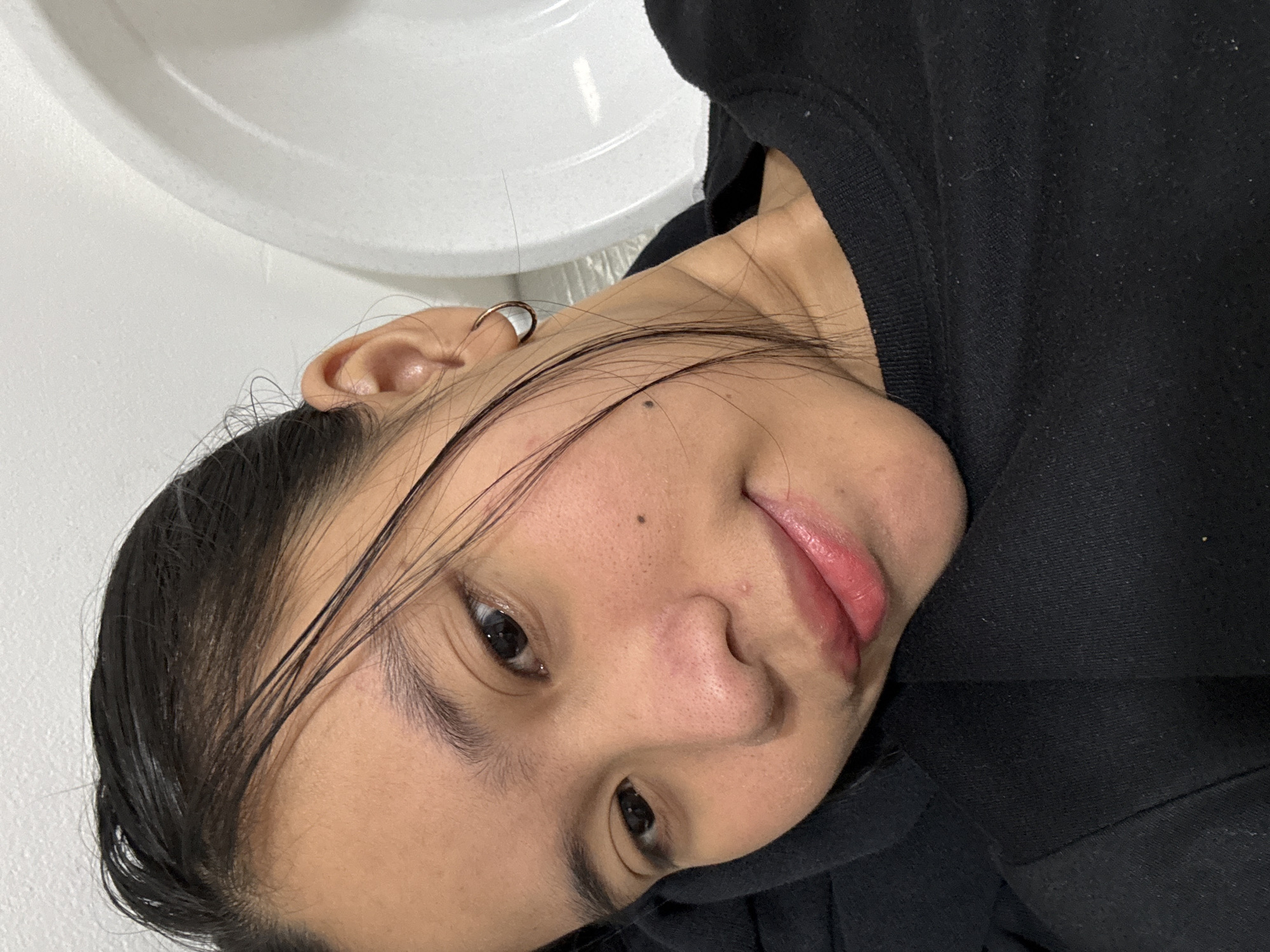 This is a close-up, chest-up, indoor selfie of a young woman with dark hair pulled back. She is wearing a black crew-neck sweatshirt and a small hoop earring in her left ear. Her head is tilted to her right, and she is looking towards the camera with a slight smile. The background appears to be a white wall and a portion of a white, curved object, possibly a sink or tub. The lighting is soft and diffused. There are no visible identifying landmarks or elements that would indicate a specific location within South Jakarta, Indonesia, or any other city.