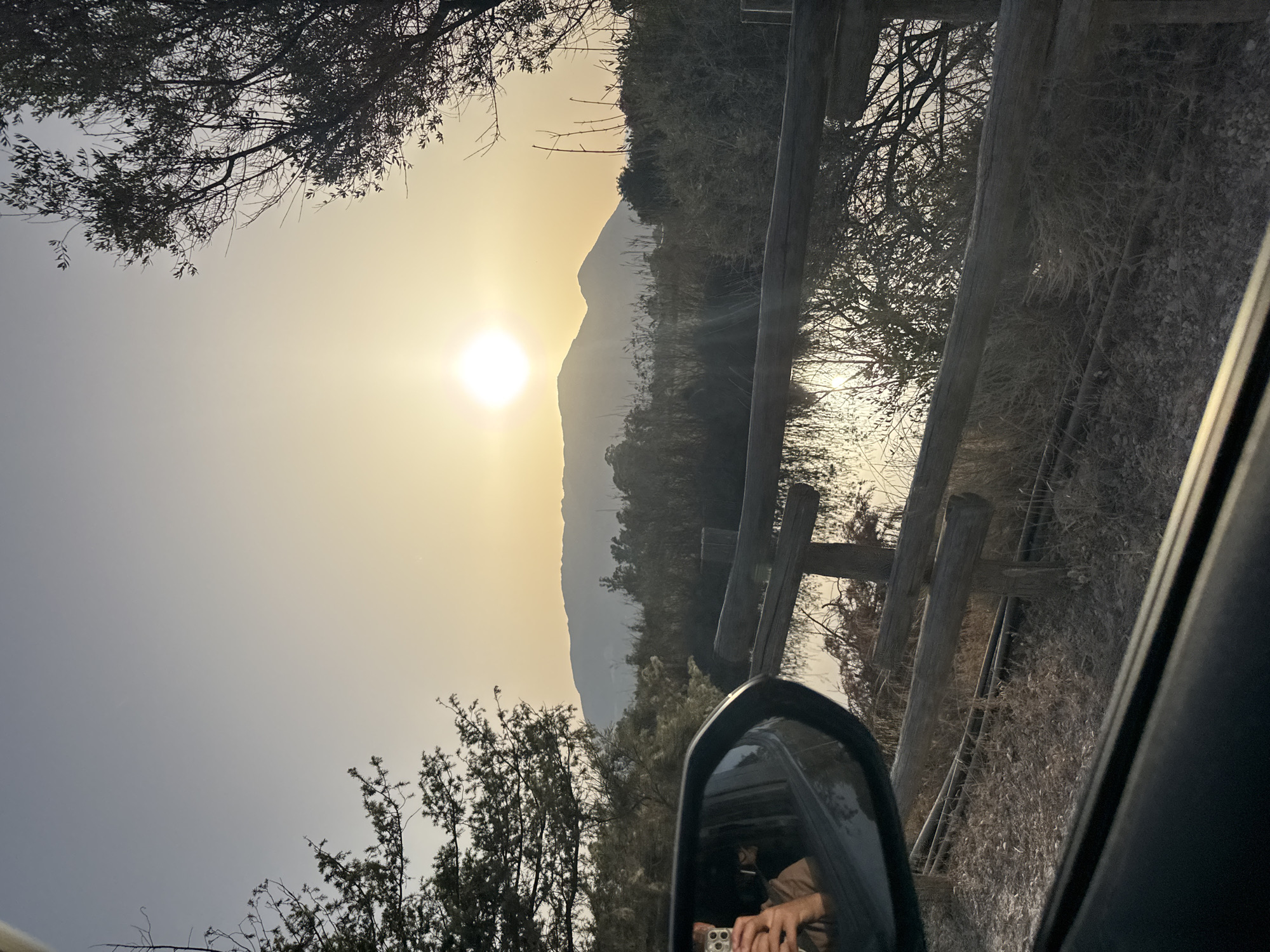 A photograph taken from inside a vehicle shows a person's reflection in the side-view mirror, holding a mobile phone. The background features a serene landscape with mountains silhouetted against a bright, hazy sunset. A rustic wooden fence runs horizontally across the mid-ground, separating a dry, grassy area from a body of water reflecting the sunlight. Trees frame the scene on the left and right, with sparse foliage contributing to the autumnal or dry season atmosphere. The overall impression is one of a tranquil, natural setting at dusk, likely near Orosei, Italy, given the mountainous terrain and Mediterranean vegetation.