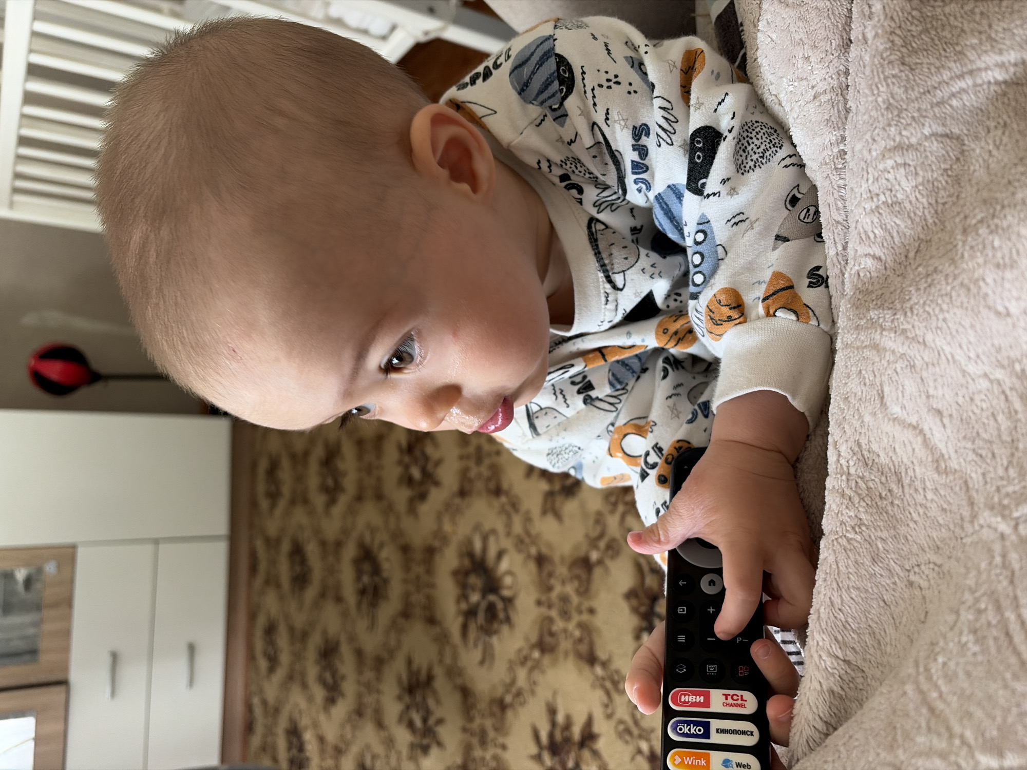A baby wearing pajamas printed with space-themed graphics is shown in a portrait-style shot. The baby is holding a black TCL TV remote control with their left hand. The background consists of a beige-patterned rug and light-colored furniture, including a crib. The image suggests an indoor environment, possibly a living room or bedroom. Based on the presence of Cyrillic alphabet labels (e.g., "иви," "ökko," "Кинопоиск") on the remote control, the location is likely in a Russian-speaking region, which is a reference to the city of Chaikovsky, Russia.