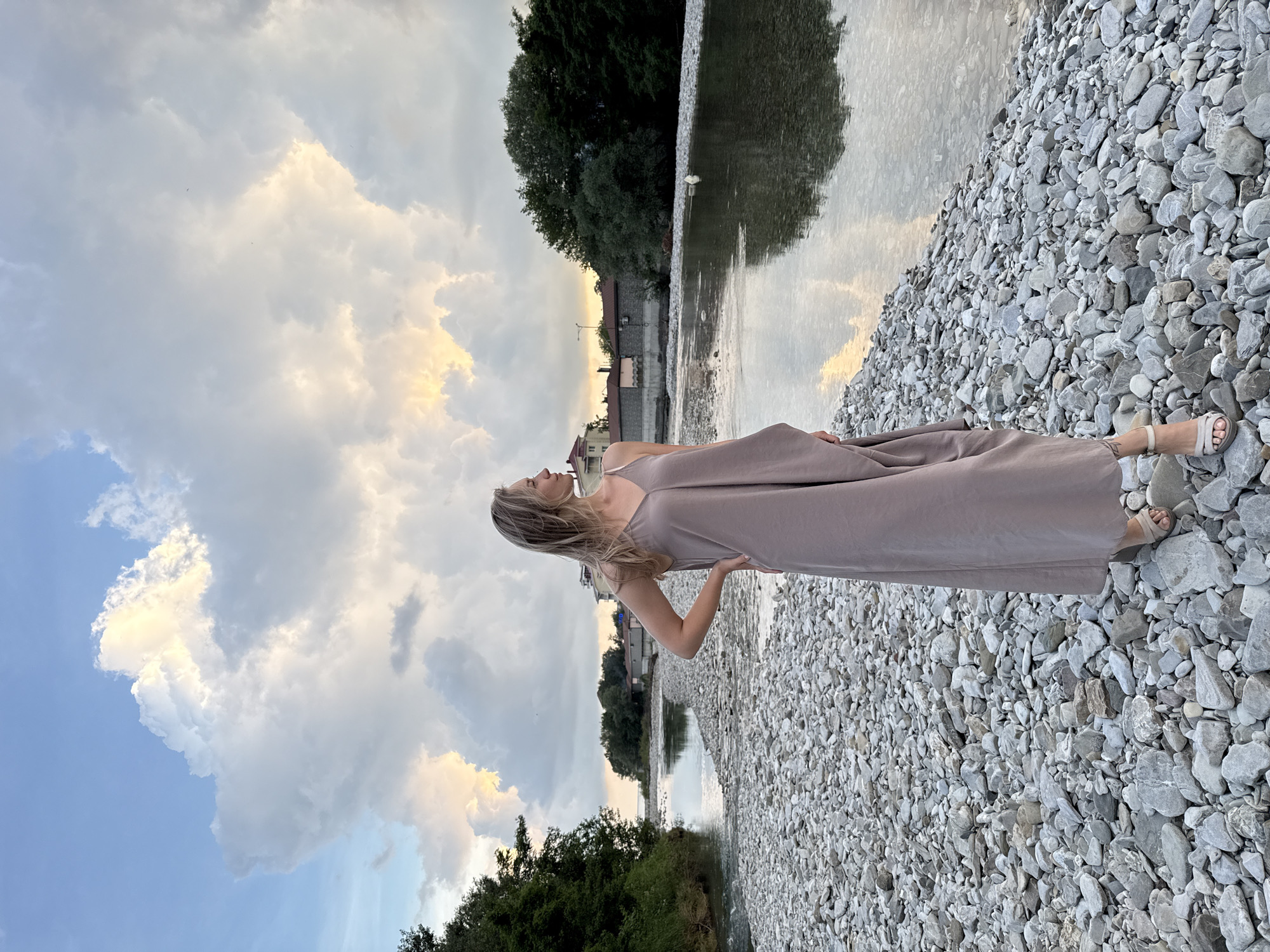 A young Caucasian woman with blonde hair stands on a rocky shore, facing a calm river. She is wearing a long, taupe-colored dress and beige sandals. Her left hand is on her hip, and she looks to her right, out towards the water. The river reflects the cloudy sky, which is illuminated by the setting sun, casting warm hues across the scene. On the opposite bank of the river, trees and buildings are visible, suggesting an inhabited area within or near the city of Nebug, Russia. The shore is covered with numerous gray and white pebbles of various sizes. The overall atmosphere is serene and tranquil.