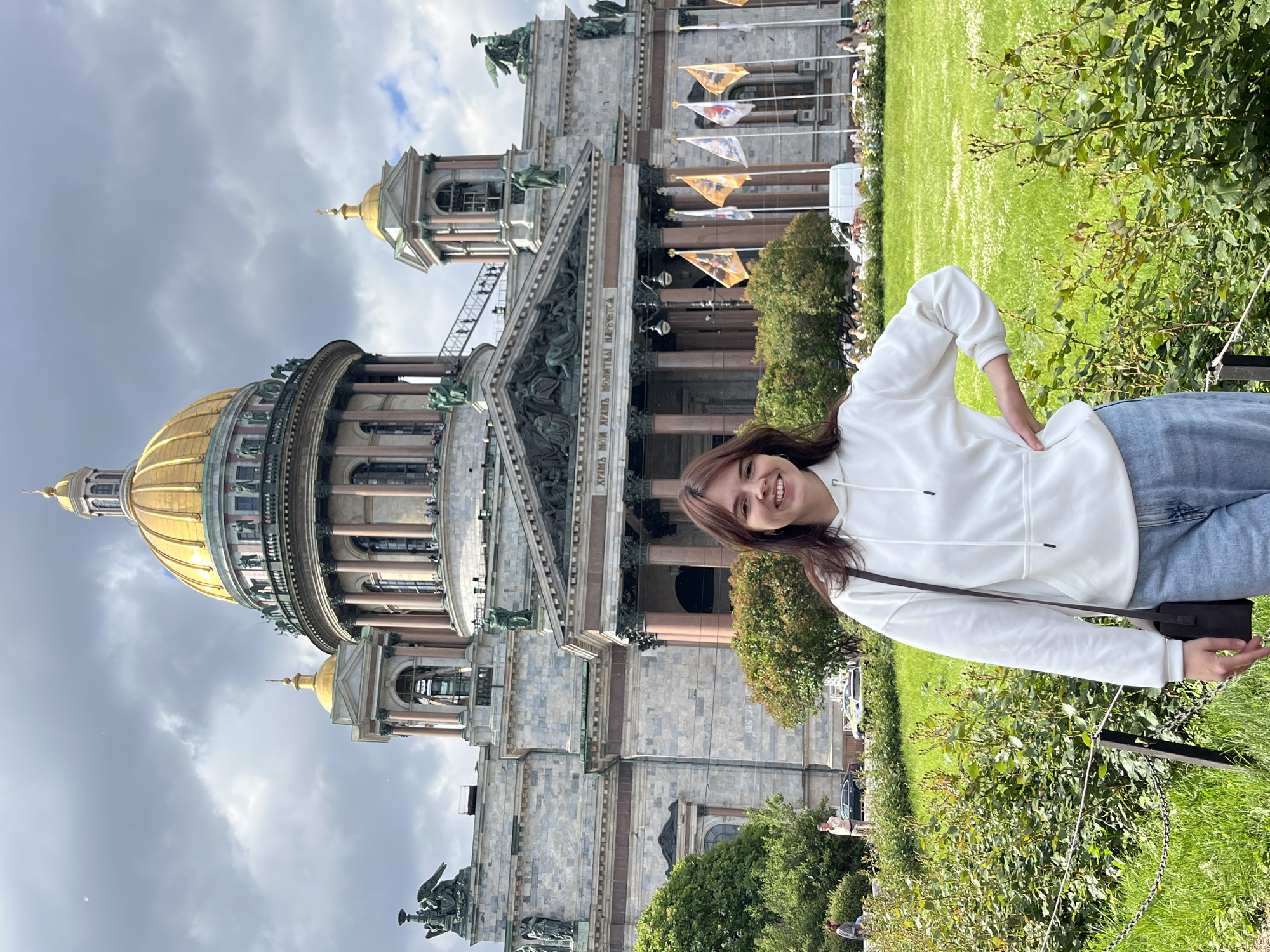 A young woman with brown hair and a white hoodie smiles at the camera, standing in front of St. Isaac's Cathedral in Saint Petersburg, Russia. She is wearing blue jeans and a dark crossbody bag. The cathedral, a prominent landmark in the city, features a large golden dome and intricate architecture. The scene is set on a partly cloudy day with lush green grass and foliage in the foreground, and other people and a white van are visible in the background.