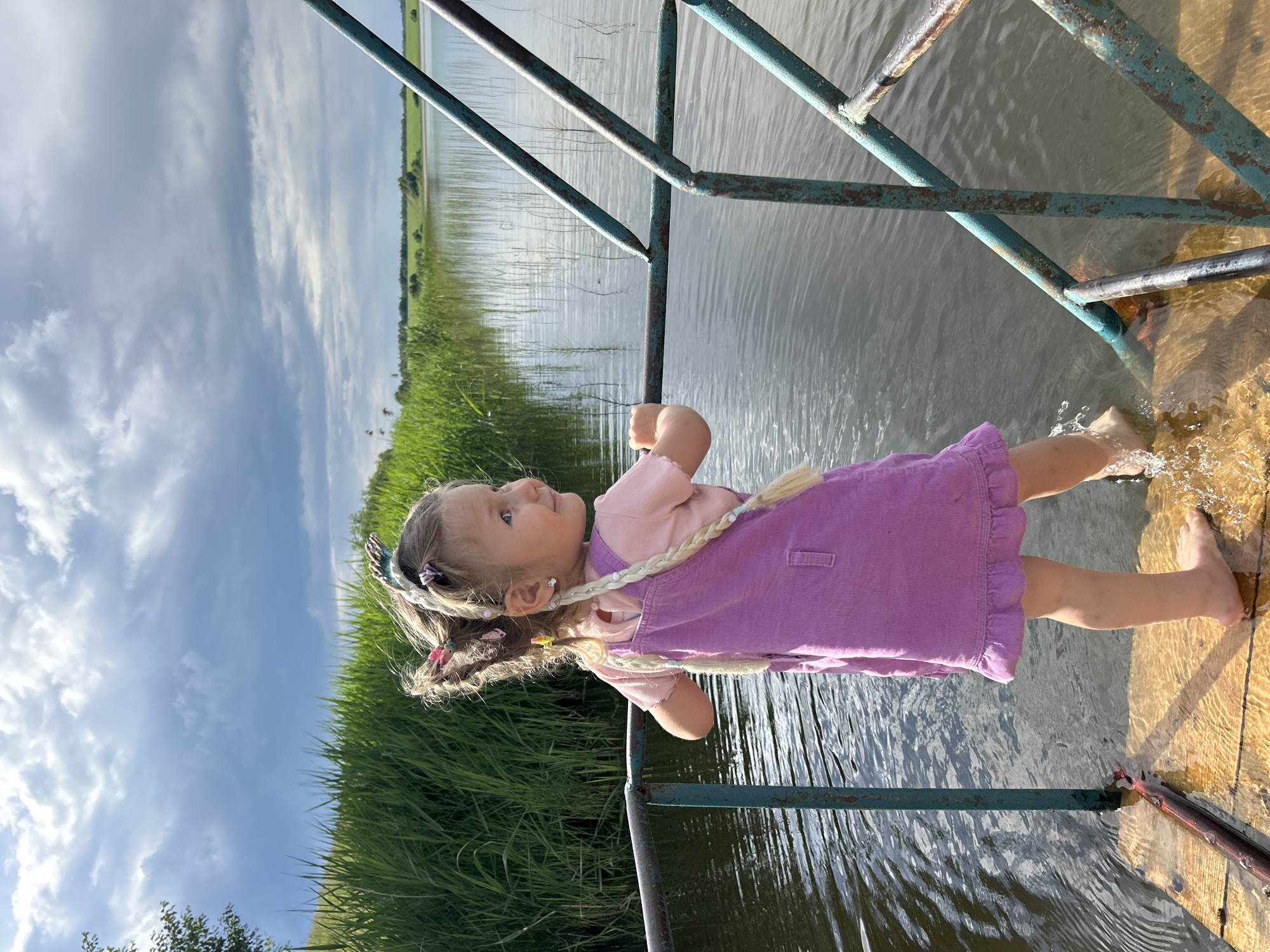 A young girl with blonde hair styled in braids, wearing a pink t-shirt and a purple pinafore dress, stands on a wooden platform next to a body of water. She is reaching her foot into the water, creating a splash. The girl is looking up and to her left with a curious expression. The background shows a vast expanse of water, reeds or tall grass along the bank, and a cloudy blue sky. A rusty metal railing separates the platform from the water. The scene appears to be outdoors during the daytime, with good lighting suggesting it could be late afternoon. The overall atmosphere is one of childhood exploration and enjoyment of nature in a rural or semi-rural setting, possibly near a lake or river in Верх-Рождество, Russia.