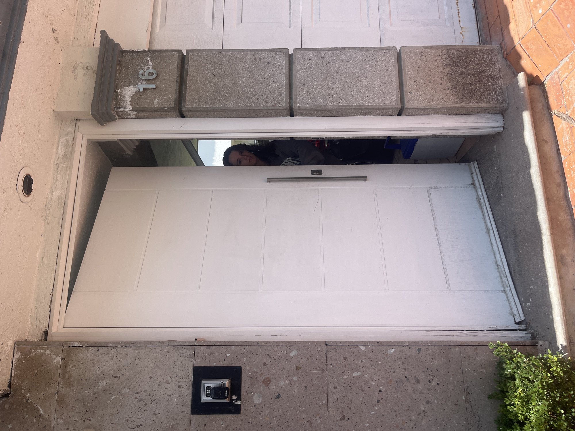 The photograph, taken from a low angle, presents the entrance to a building. The door is white with recessed panel detailing and a horizontal metal handle. Above the door is a decorative stone lintel with the number "16" affixed to the left side. A person is visible through the partially open doorway, appearing to look out. To the right of the doorway, a small green bush is visible. A security panel is mounted on the lower stone wall to the right. The surrounding area consists of various masonry and concrete surfaces. The scene appears to be located in San Bernardino Tlaxcalancingo, Mexico.