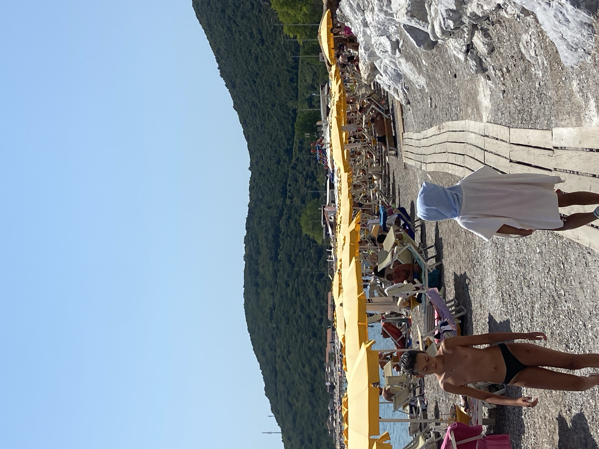 This is a photograph of a crowded beach with numerous yellow umbrellas and lounge chairs. The beach is situated in Prijevor, Montenegro, with a large, tree-covered mountain serving as a backdrop. The sky is clear and blue. In the foreground, two children are walking on a wooden pathway. The child on the left, a boy wearing black swim trunks, is facing forward, looking down. The child on the right, wearing a white cover-up with a light blue hood, is also facing forward and walking away from the camera. The beach appears to be made of pebbles and sand.