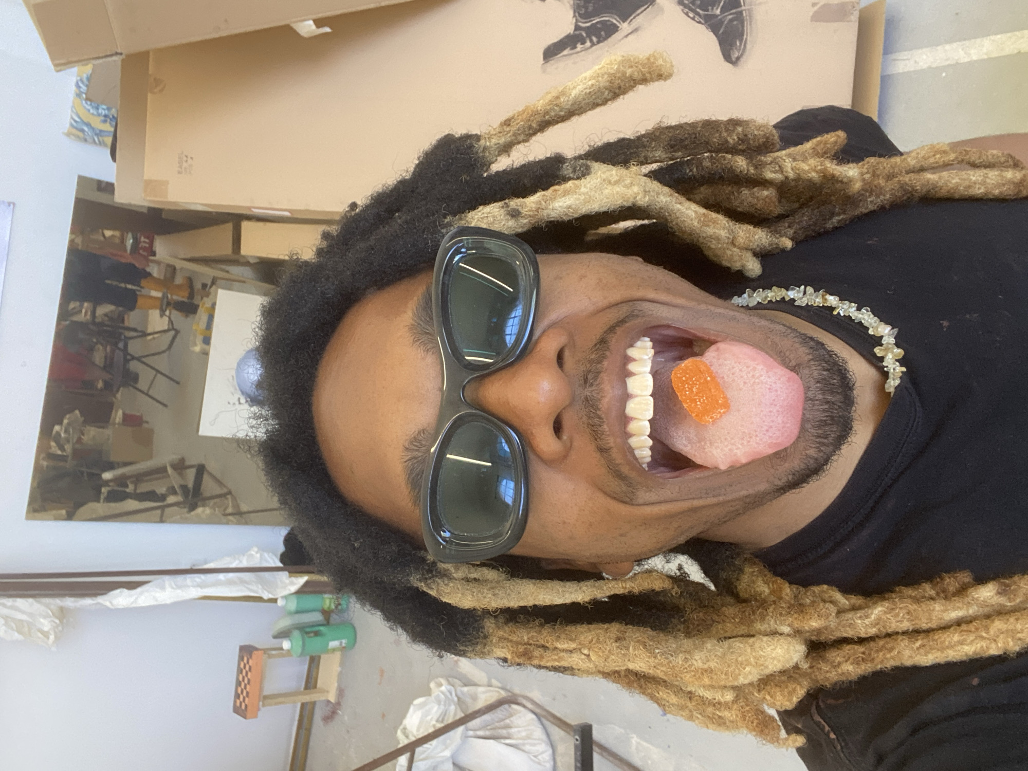 The image is a close-up self-portrait of a person with dark skin, long dreadlocks (some dark, some blonde/light brown), wearing black sunglasses and a black t-shirt. A necklace made of small, irregularly shaped, light-colored beads is visible around their neck. The person's mouth is wide open, with their tongue fully extended, and an orange, rectangular gummy candy rests on the tip of their tongue. They have a short beard or stubble.
The setting appears to be an art studio or workshop. In the background, to the left of the person's head, there is a large mirror reflecting parts of a room with furniture, including chairs and tables. Above the person's head, a large cardboard box is visible, with faint text "EAGLE" and numbers "09/14" on it. Behind the box, a piece of artwork depicting parts of legs and shoes can be seen. To the right of the person's body, elements like a small wooden stand with a checkered pattern and some green bottles (possibly paint or art supplies) are present. The reflections in the sunglasses show horizontal lines, possibly from light fixtures or windows.
The activity is a playful pose for the camera, showcasing the gummy candy on the tongue. The overall emotion conveyed is lighthearted and quirky. No specific time of day or weather is discernible. The general ambiance suggests an indoor, creative environment.