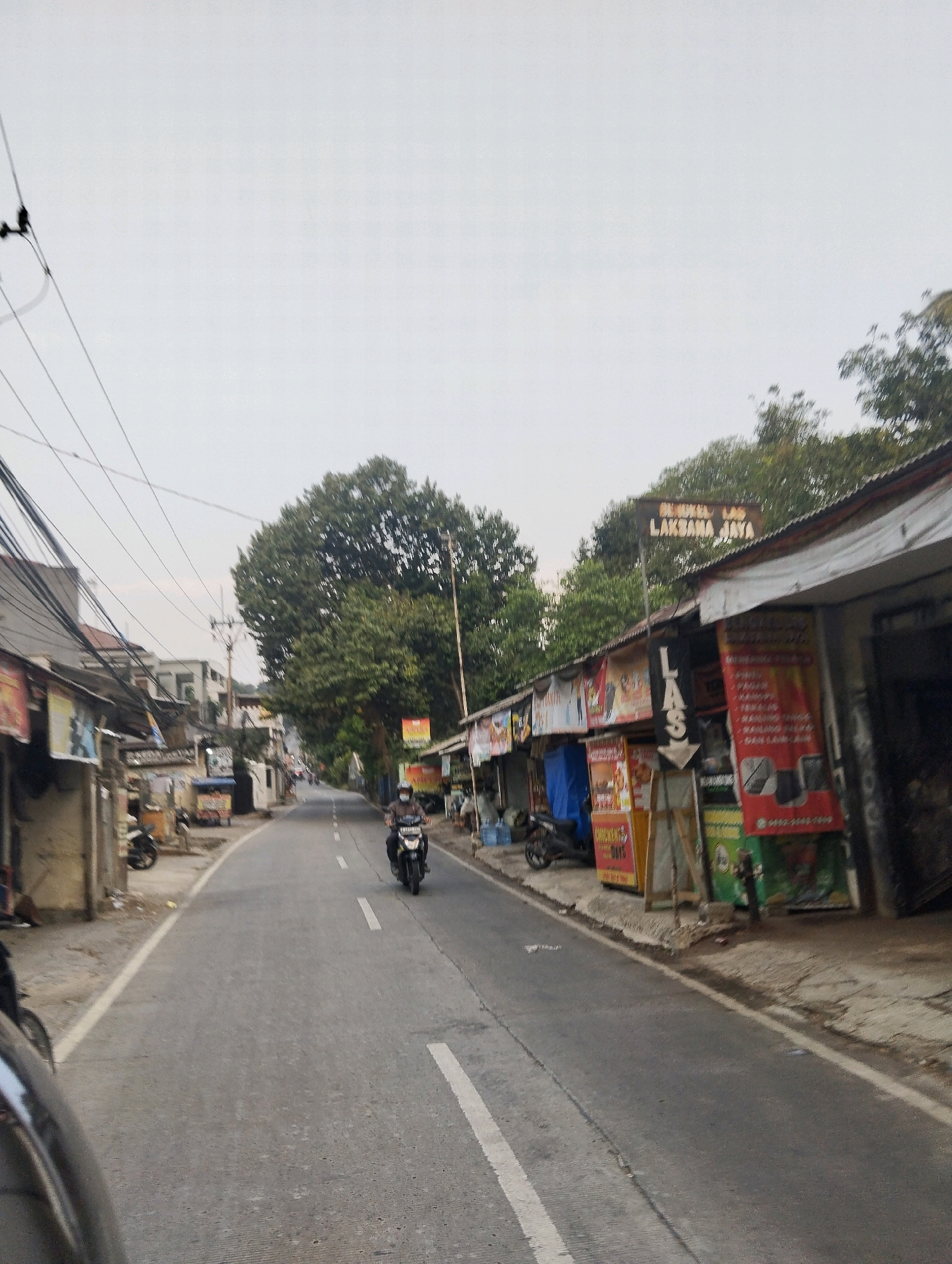 The image captures a street scene in Megamendung, Indonesia, characterized by a paved road lined with shops and lush trees. The road itself has white markings dividing the lanes, and a motorcycle rider is seen heading away from the camera, wearing a mask and helmet. The shops on the right side of the road display various banners and signs, including one that reads "LAKSAMA JAYA." One banner features text that appears to be in Indonesian, with words like "MURAH" (cheap) and "MAHAL" (expensive), and possibly product descriptions or prices related to electronics. Another banner has a phone number and images of a phone and laptop. To the left of the road, more shops and buildings are visible, along with parked motorcycles. Overhead, power lines crisscross the sky, and dense foliage from trees forms a canopy over parts of the street and shops. The sky is overcast, suggesting a diffused light source.